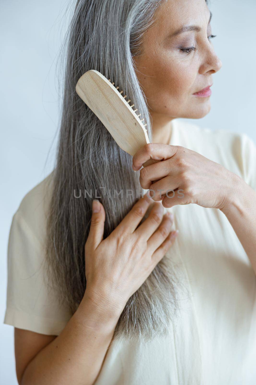 Tranquil middle aged Asian model brushes long hoary hair posing on light background in studio. Mature beauty lifestyle