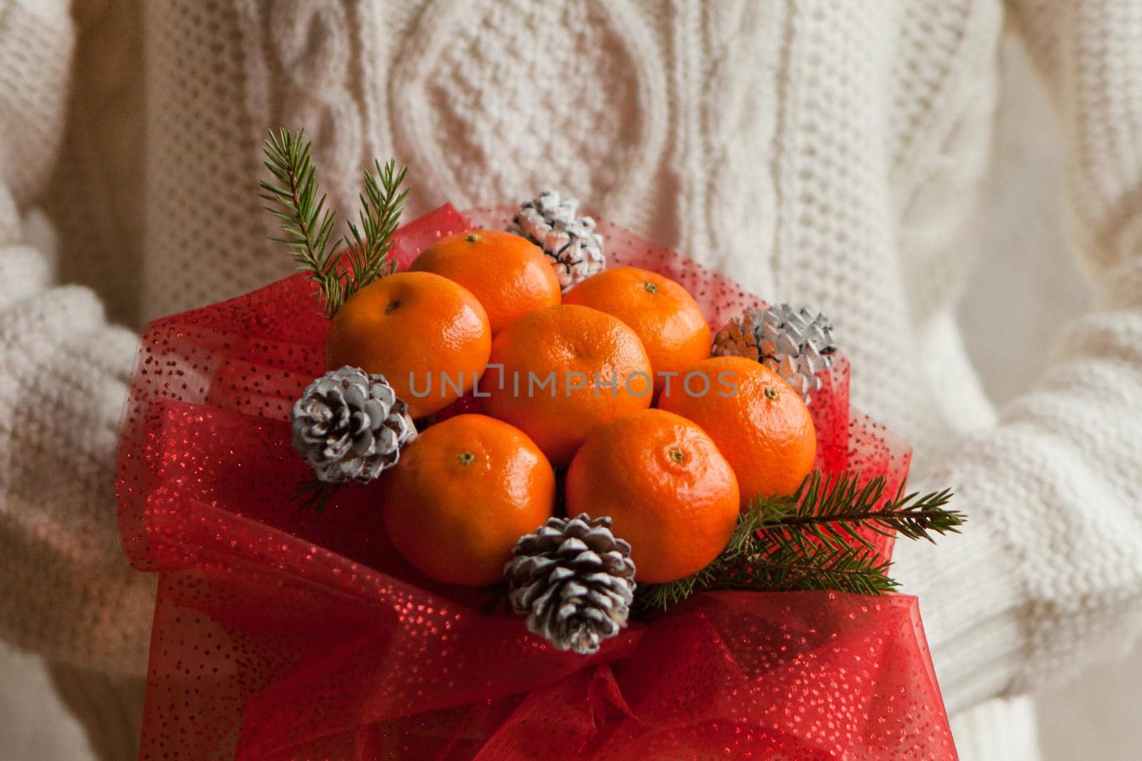 Woman hands in white sweater holding bouquet of mandarins and Christmas tree branches. New Year's edible bouquet of fruits. Gift for Christmas. useful gift made of fruits. decor of fruits.