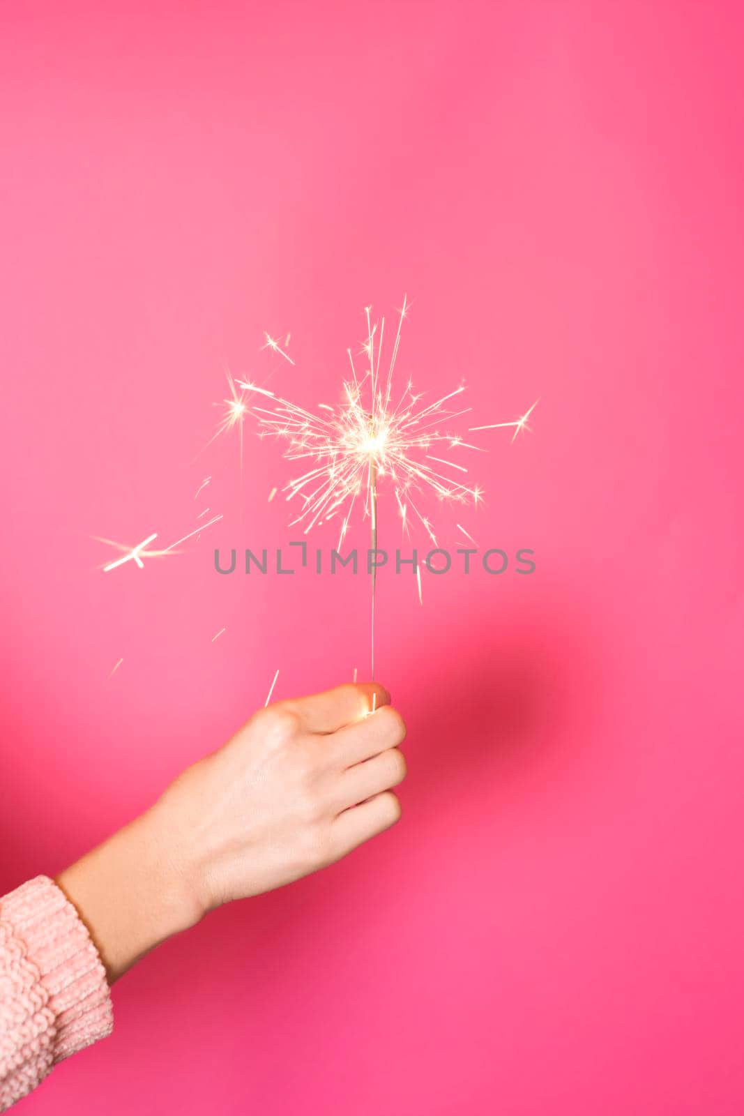 Close up of female hand with burning Christmas sparkler on pink background. Happy New Year celebration concept