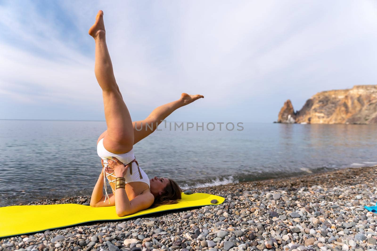 Young woman in white swimsuit with long hair practicing stretching outdoors on yoga mat by the sea on a sunny day. Women's yoga fitness pilates routine. Healthy lifestyle and meditation concept by panophotograph
