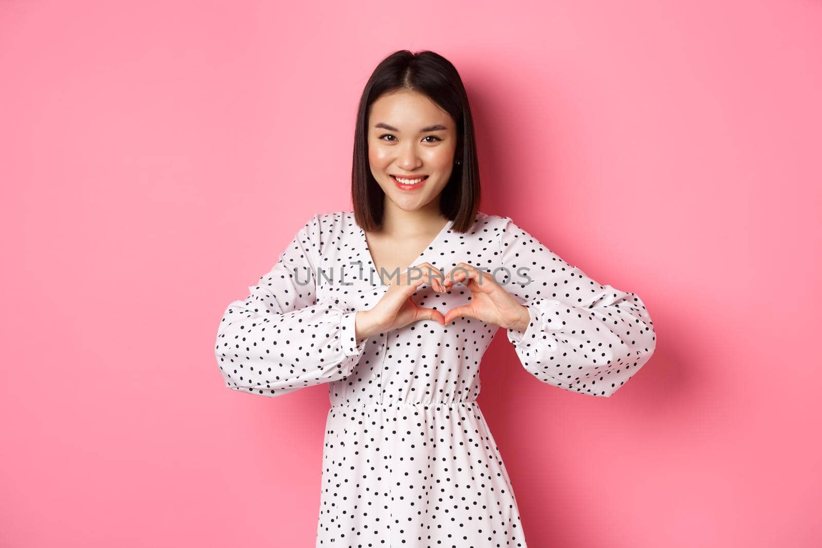 Romantic asian woman showing heart sign, I love you gesture, smiling cute at camera, standing in dress over pink background.