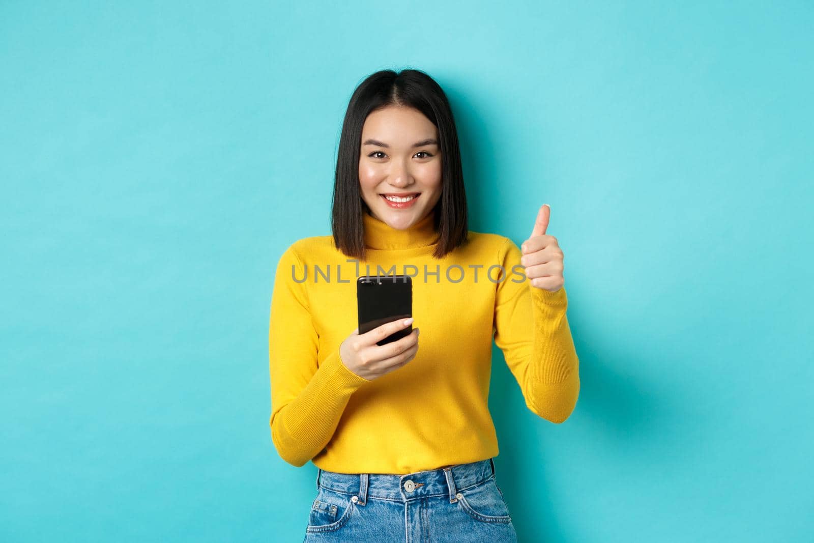 E-commerce and online shopping concept. Satisfied asian female client showing thumbs up and using smartphone, smiling pleased at camera, blue background by Benzoix