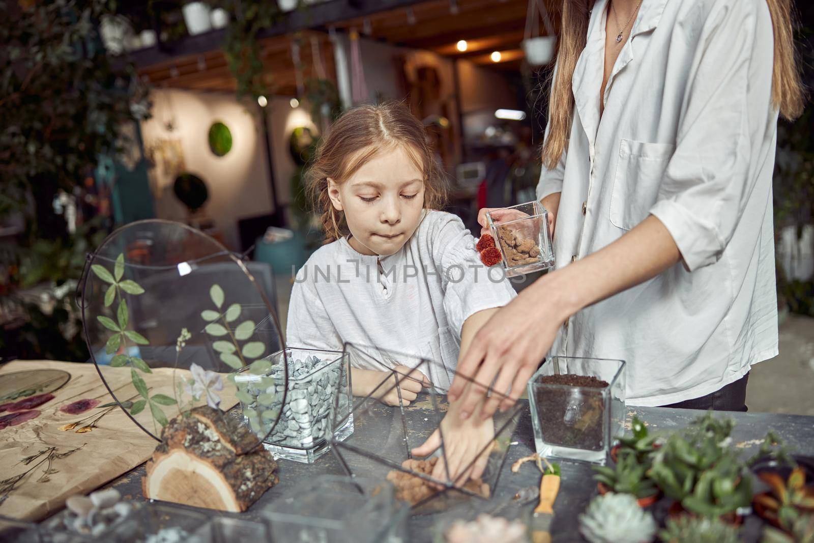 caucasian confident happy florist is working with her young daughter and making composition from glass stones and plants in botanic shop