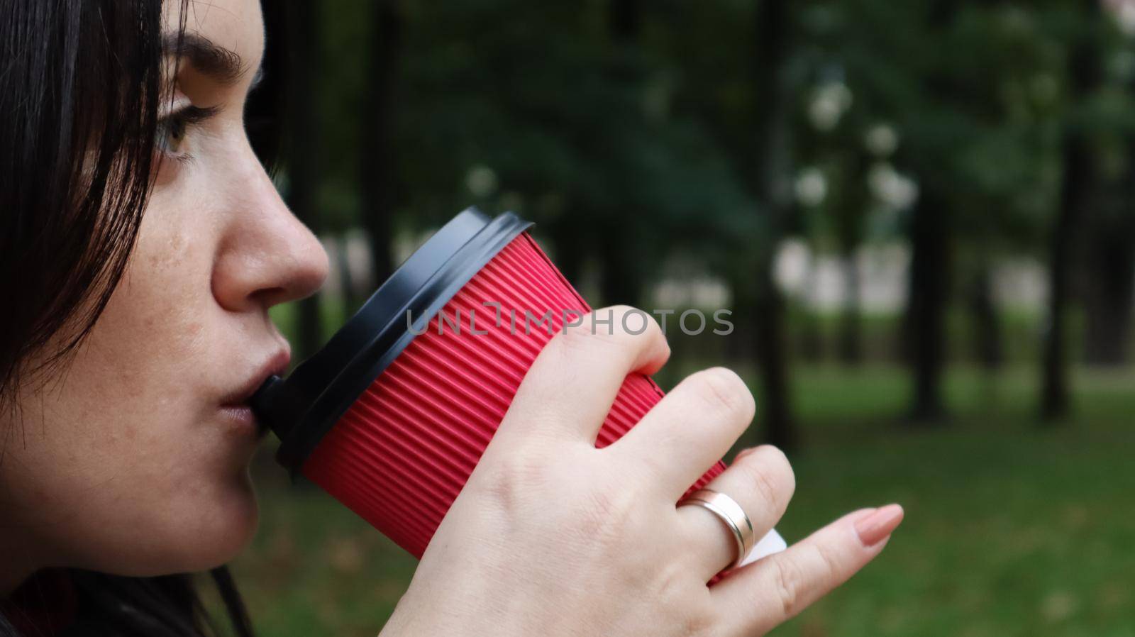 Young stylish woman drinks morning hot coffee in red eco paper cup outdoors in autumn park. Close-up side view of young woman holding takeaway coffee cup. by Roshchyn