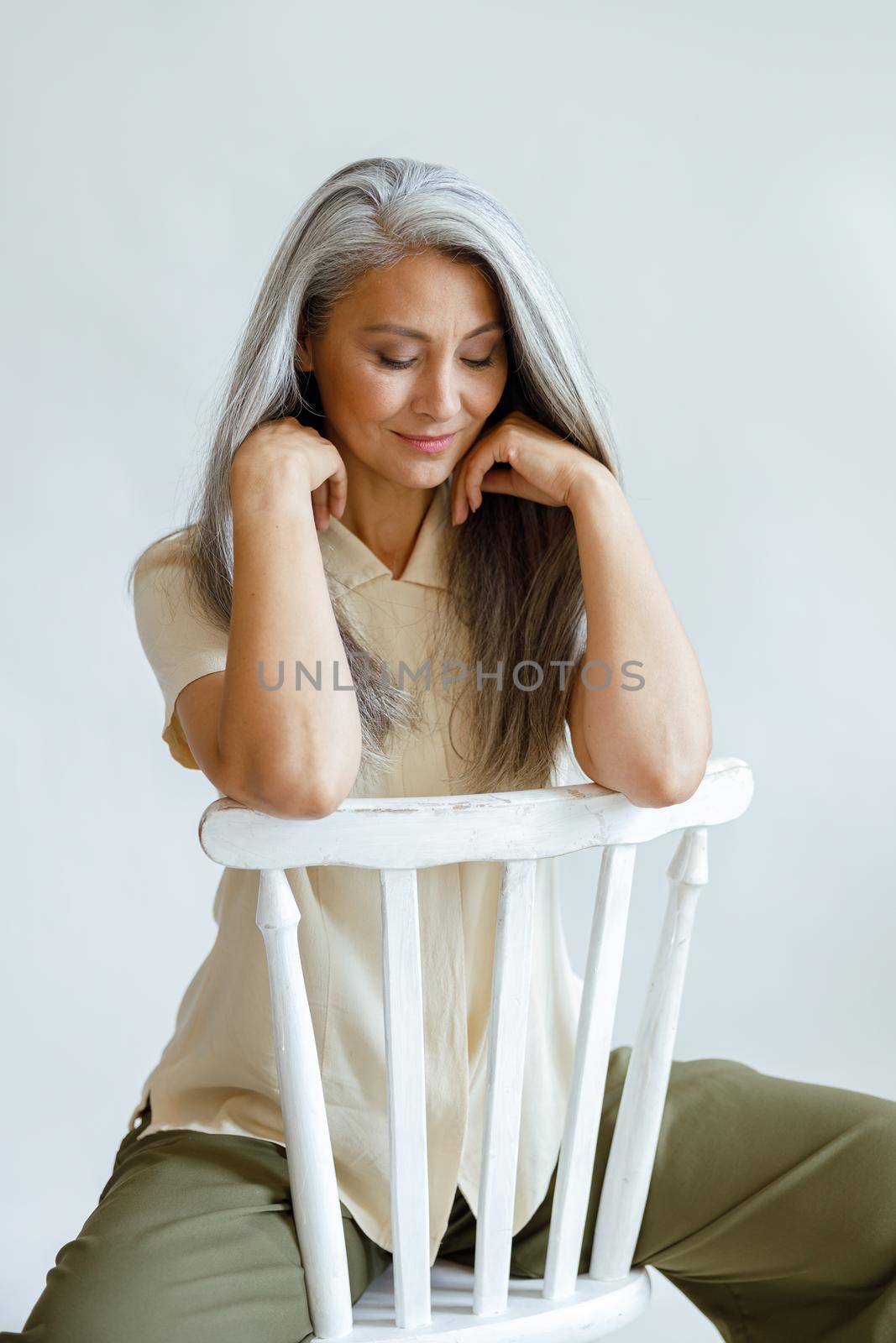 Attractive middle aged Asian woman with grey hair sits backwards on chair on light background in studio. Mature beauty lifestyle