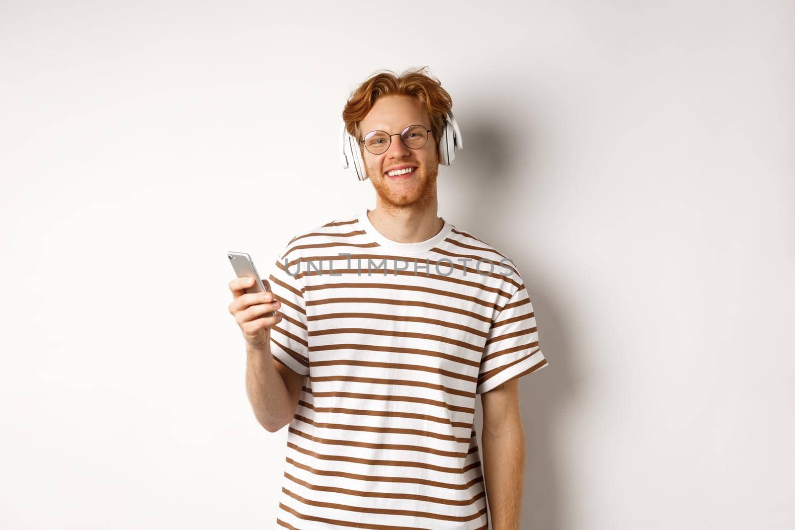 Technology concept. Young man with red hair and beard listening music in headphones and using smartphone, smiling at camera, white background by Benzoix