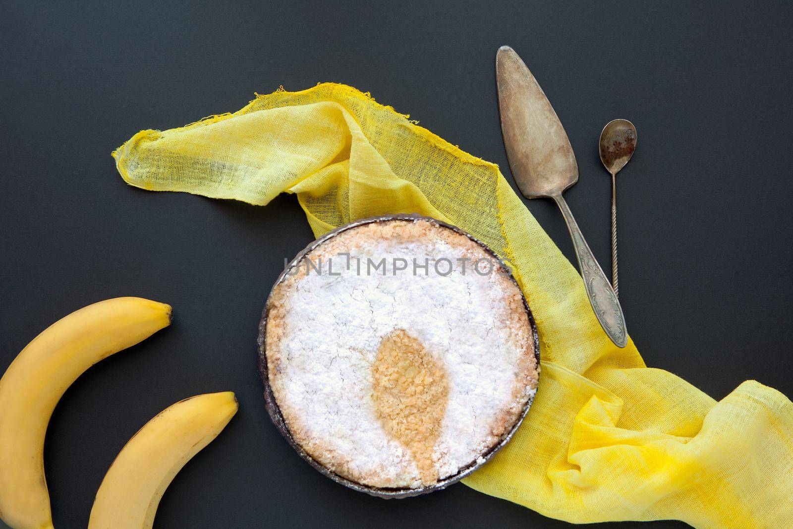 Banana cake on a black background with yellow blanket and retrospoons. Homemade pie on a dark backdrop and powdered sugar spoon shadow.