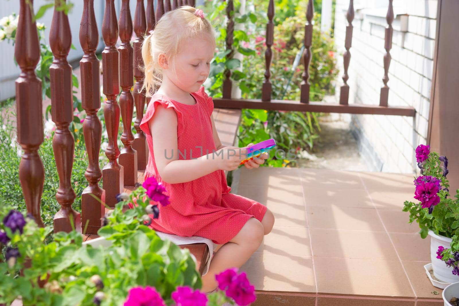 Cute caucasian fair hair girl in pink dress is playing and showing funny trendy silicone antistress colorful toy popit. Push Popper fidget toy is the ideal toy to get rid of your frustration or stress.