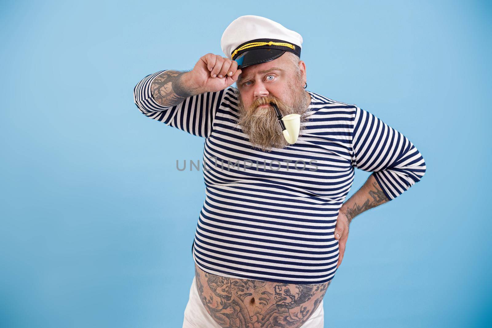 Mature fat sailor with beard and vintage smoking pipe adjusts hat looks into camera on light blue background in studio