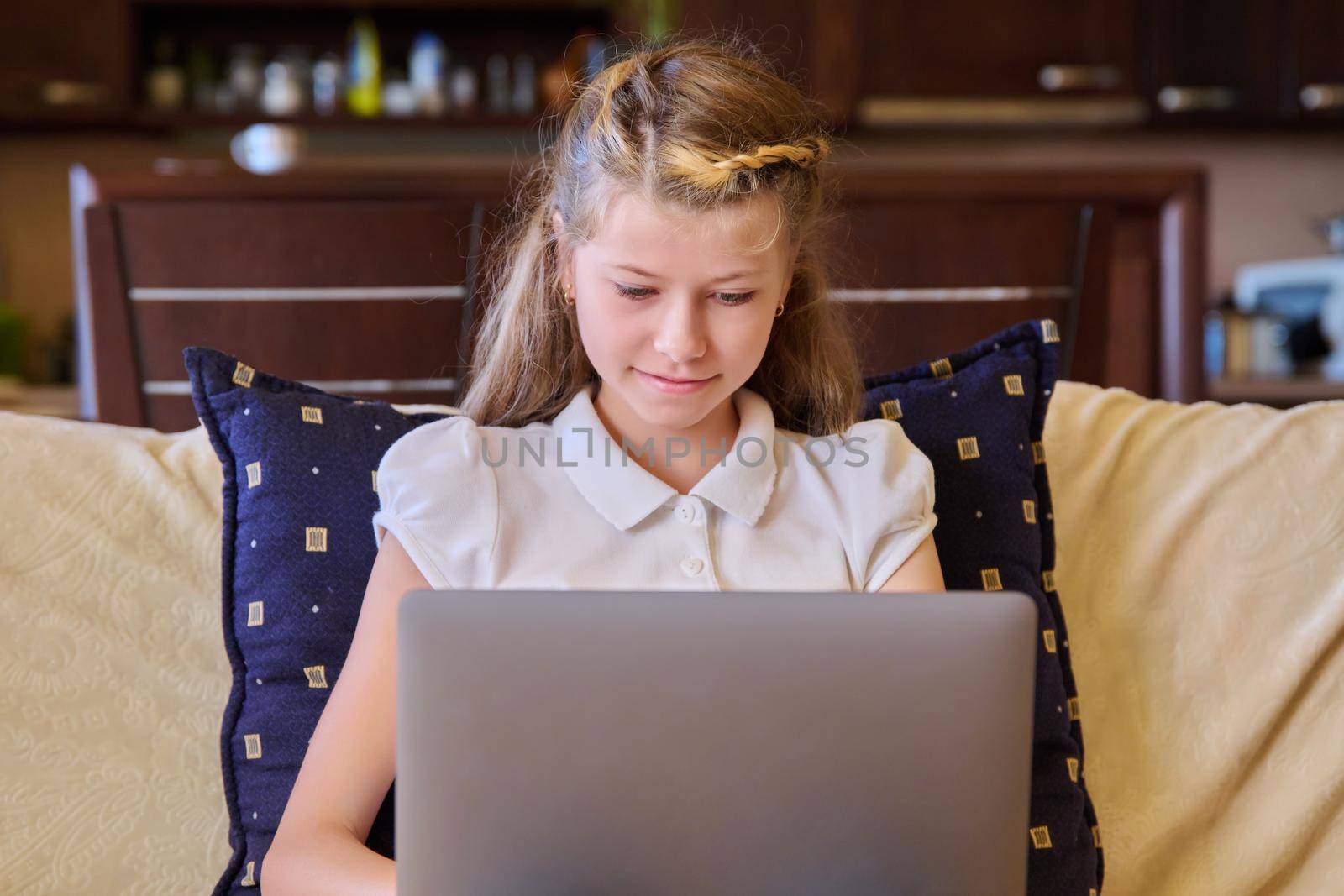 Child girl with laptop at home on the couch. Preteen blonde girl looking at laptop screen, education, relaxation, leisure, computer game, video communication, home lifestyle