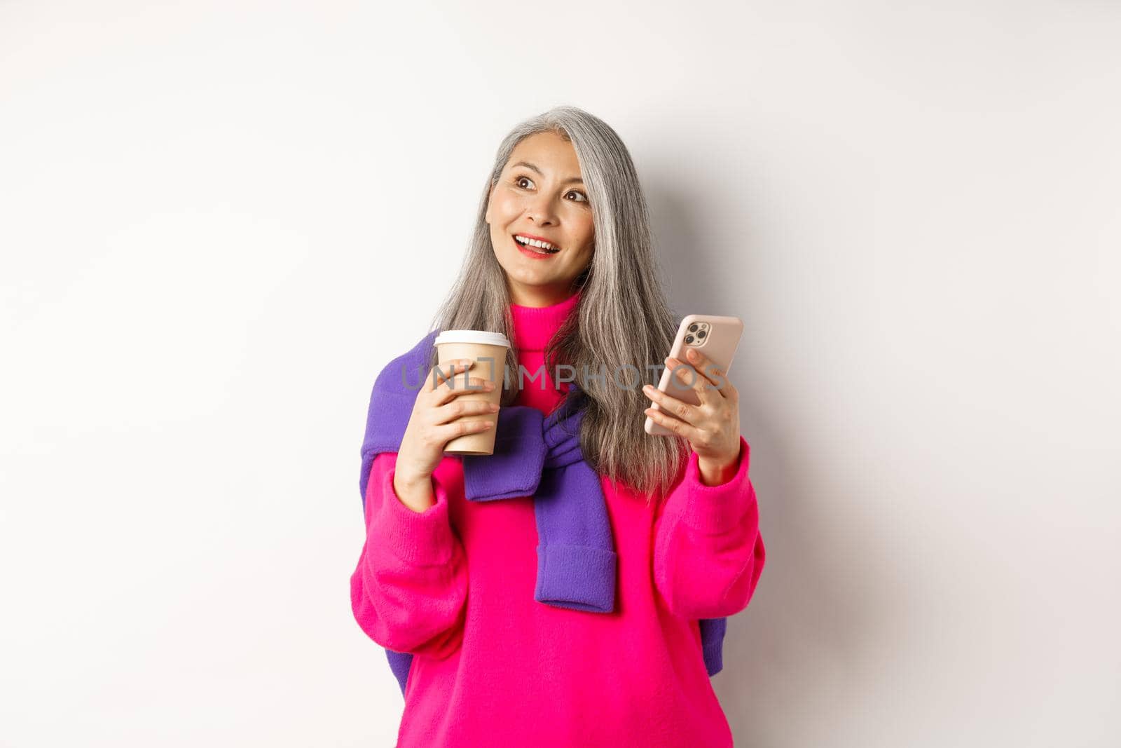 Fashionable asian mature woman with grey hair, drinking coffee and using smartphone, looking upper left corner promotion with dreamy face, white background.