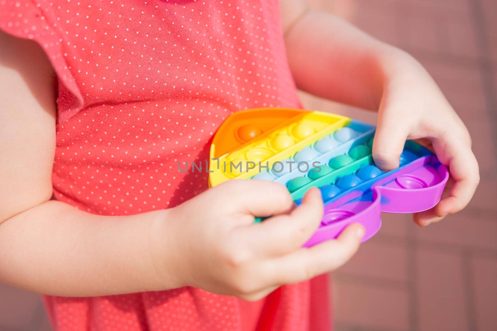 Children's hands hold a silicone toy. Push Popper fidget toy is the ideal toy to get rid of your frustration or stress. Kid having fun with popular simple dimple multicolored rainbow colors toy. Kids being kids LGBTQ colors Vertical. Summer vibes. Outdoor Outside.