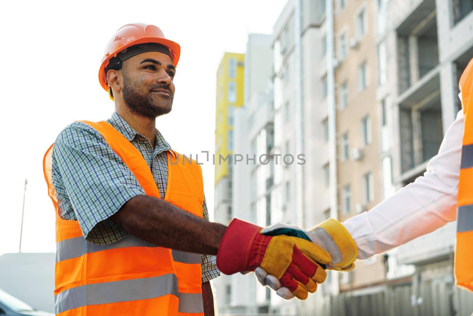 Two men engineers in workwear shaking hands against construction site, close up