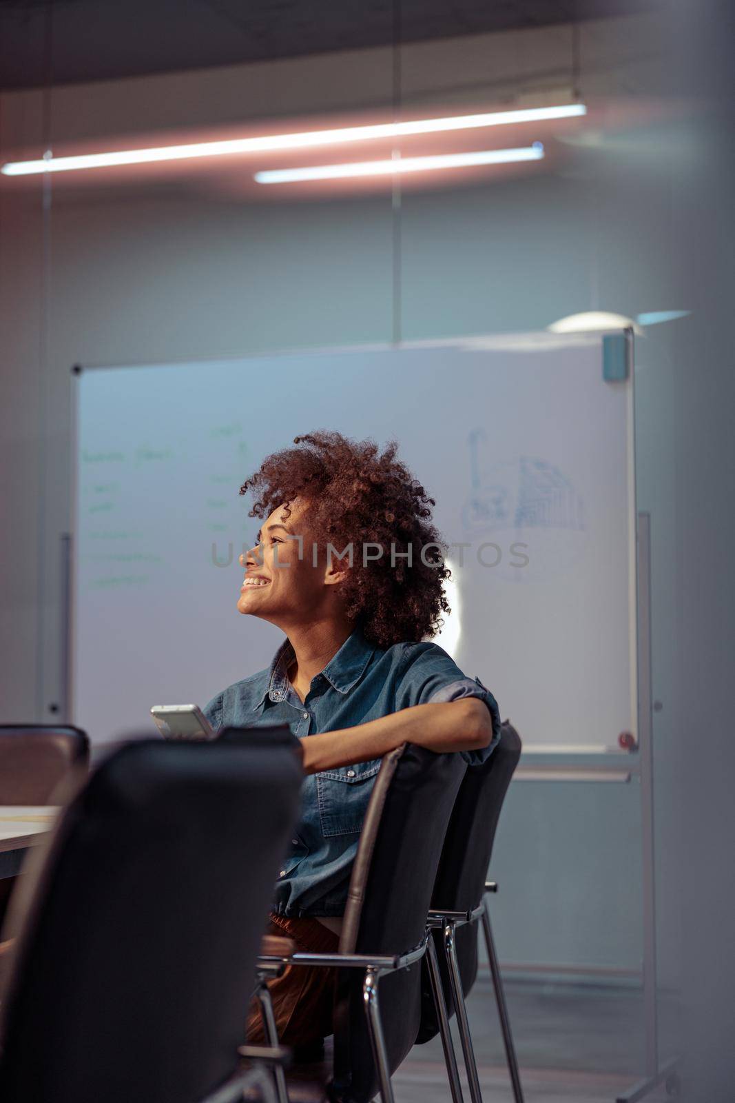 Happy female worker sitting at the table in the meeting room and holding mobile phone