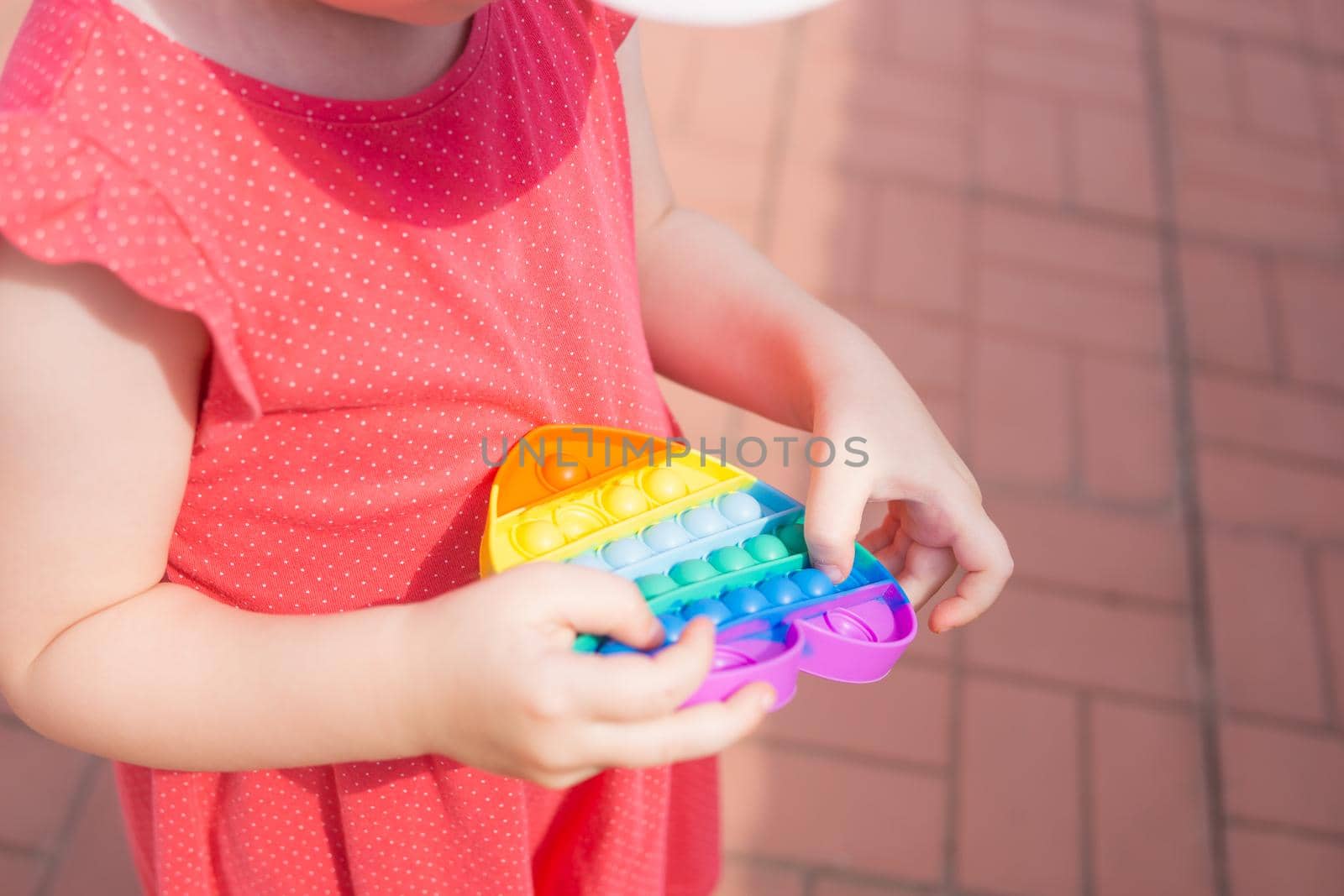 Cute caucasian fair hair girl in pink dress is playing and showing funny trendy silicone antistress colorful toy popit. Kid having fun with popular simple dimple multicolored rainbow colors toy. Children's hands hold a silicone toy. Kids being kids LGBTQ colors Vertical.