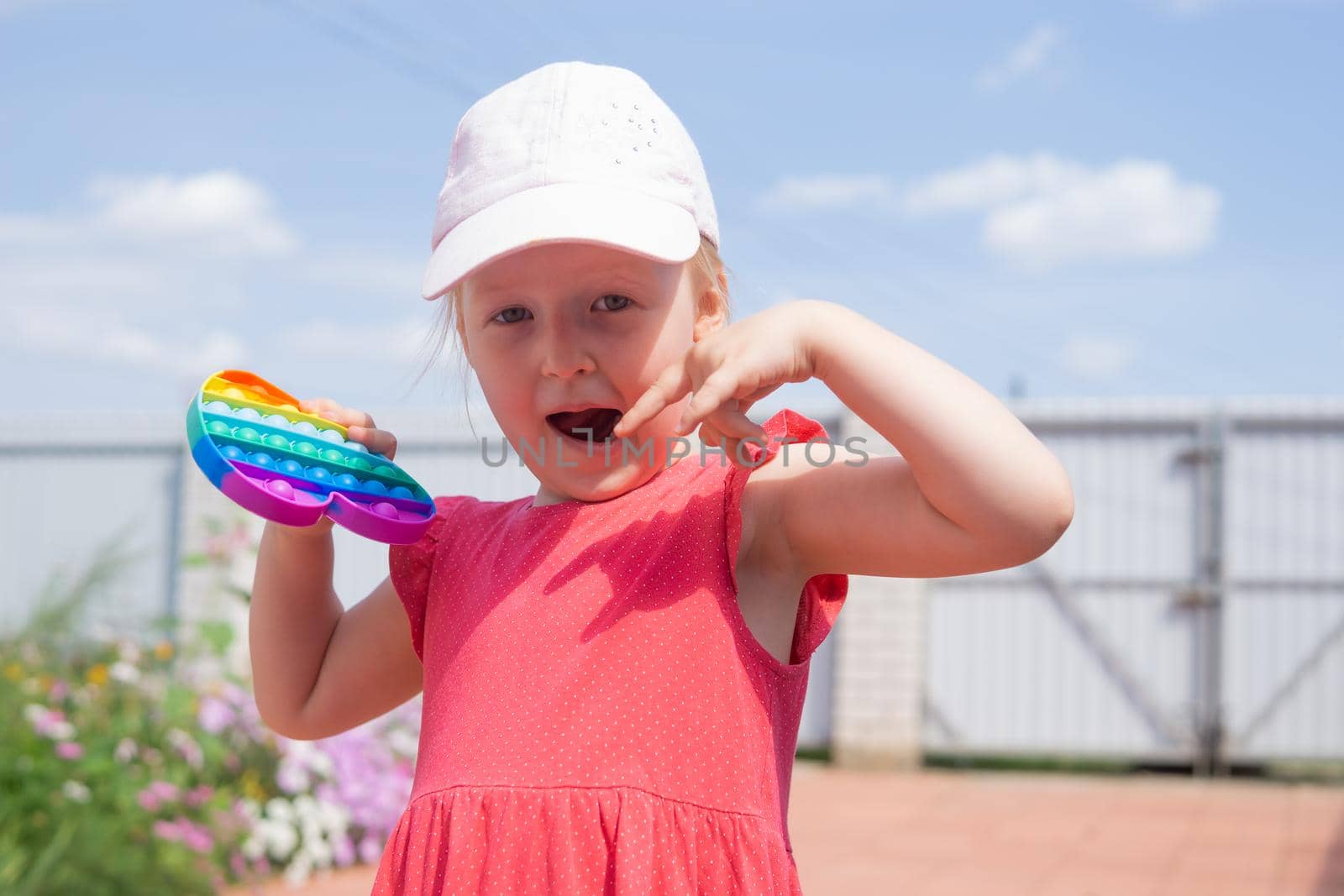 Cute caucasian fair hair girl in pink dress is playing and showing funny trendy silicone antistress colorful toy popit. Push Popper fidget toy is the ideal toy to get rid of your frustration or stress.