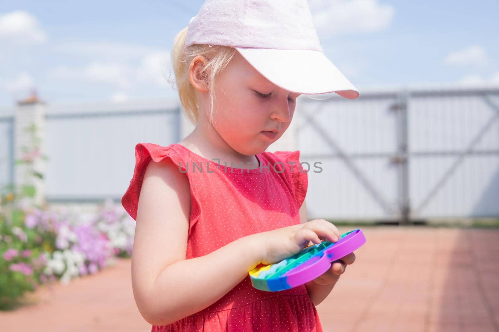 Cute caucasian fair hair girl in pink dress is playing and showing funny trendy silicone antistress colorful toy popit. Push Popper fidget toy is the ideal toy to get rid of your frustration or stress.