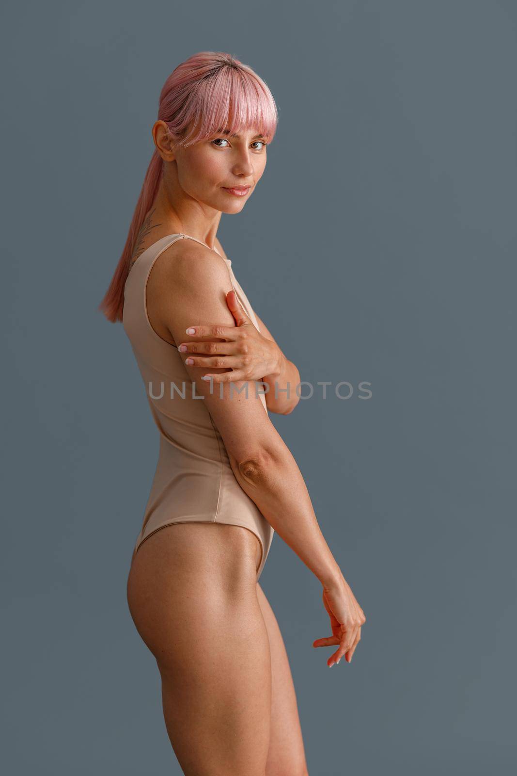 Attractive young woman with pink hair in beige bodysuit looking at camera, standing isolated over gray studio background by Yaroslav_astakhov
