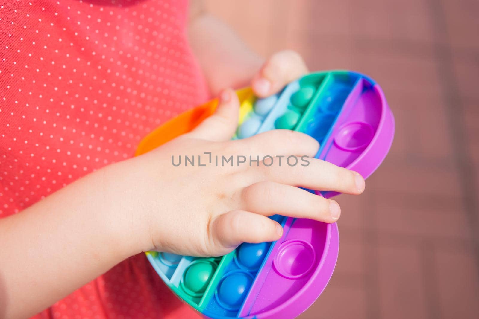Cute caucasian fair hair girl in pink dress is playing and showing funny trendy silicone antistress colorful toy popit. Kid having fun with popular simple dimple multicolored rainbow colors toy. Children's hands hold a silicone toy. Kids being kids LGBTQ colors Vertical.