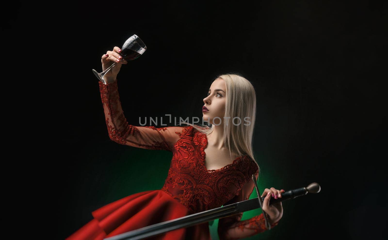 beautiful girl in a red dress with a sword and a glass of wine posing on a black background in the Studio ( blonde )