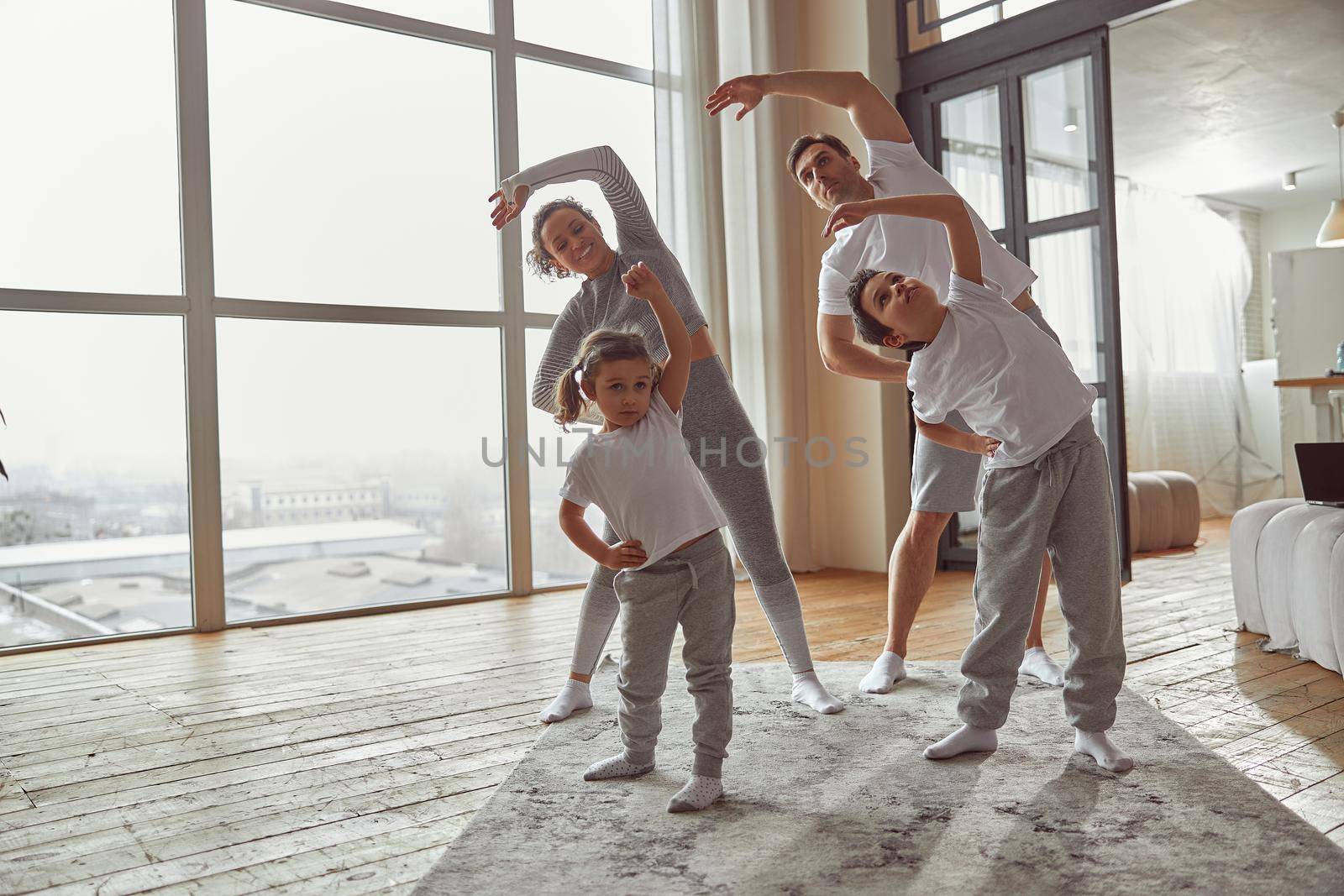 Merry mom and dad are exercising with their daughter and son near large window in living room