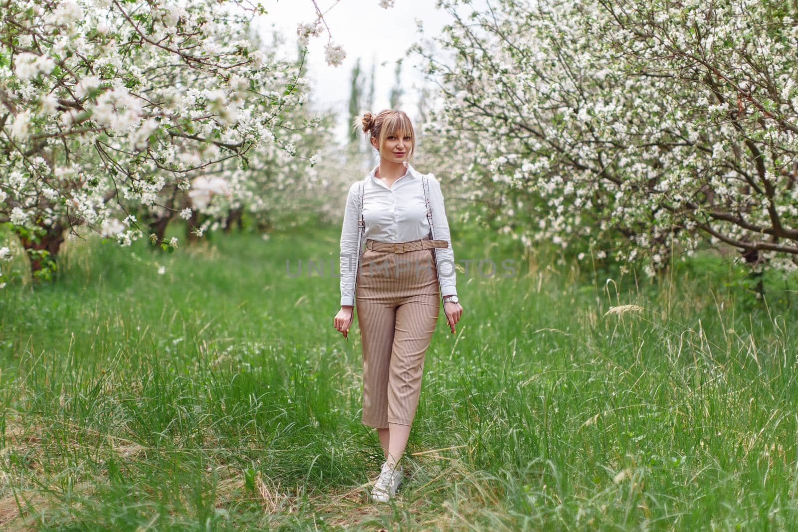 Beautiful young blonde woman in white shirt with backpack posing under apple tree in blossom in Spring garden by OnPhotoUa