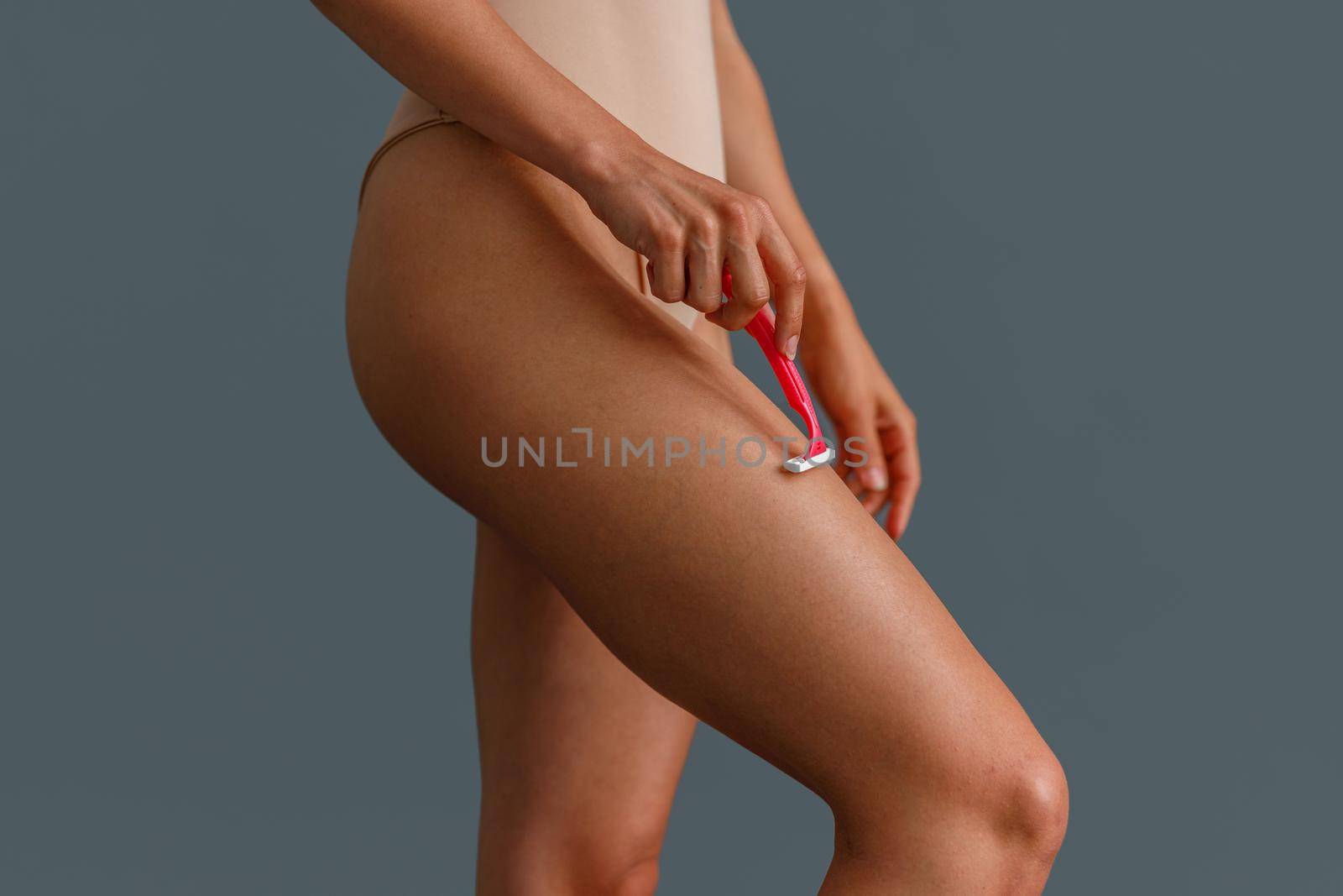 Close up shot of woman shaving her leg using razor, standing over gray studio background. Beauty, body care concept