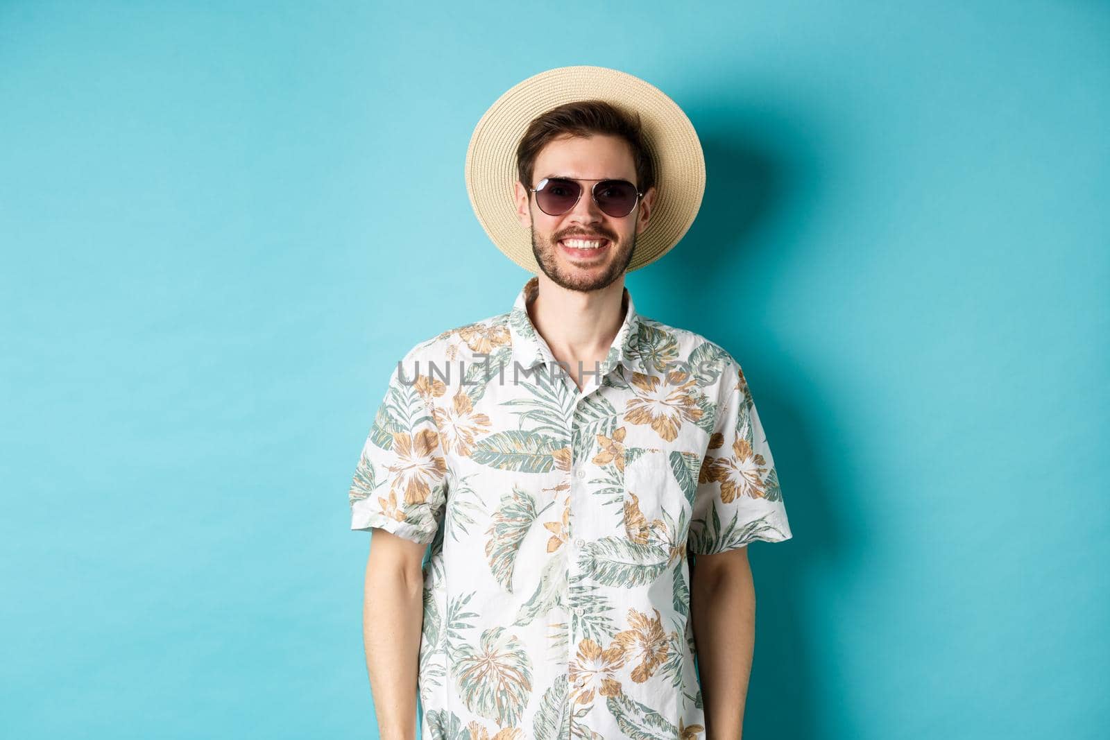 Handsome smiling guy in sunglasses and summer shirt, enjoying vacation on tour, standing on blue background.