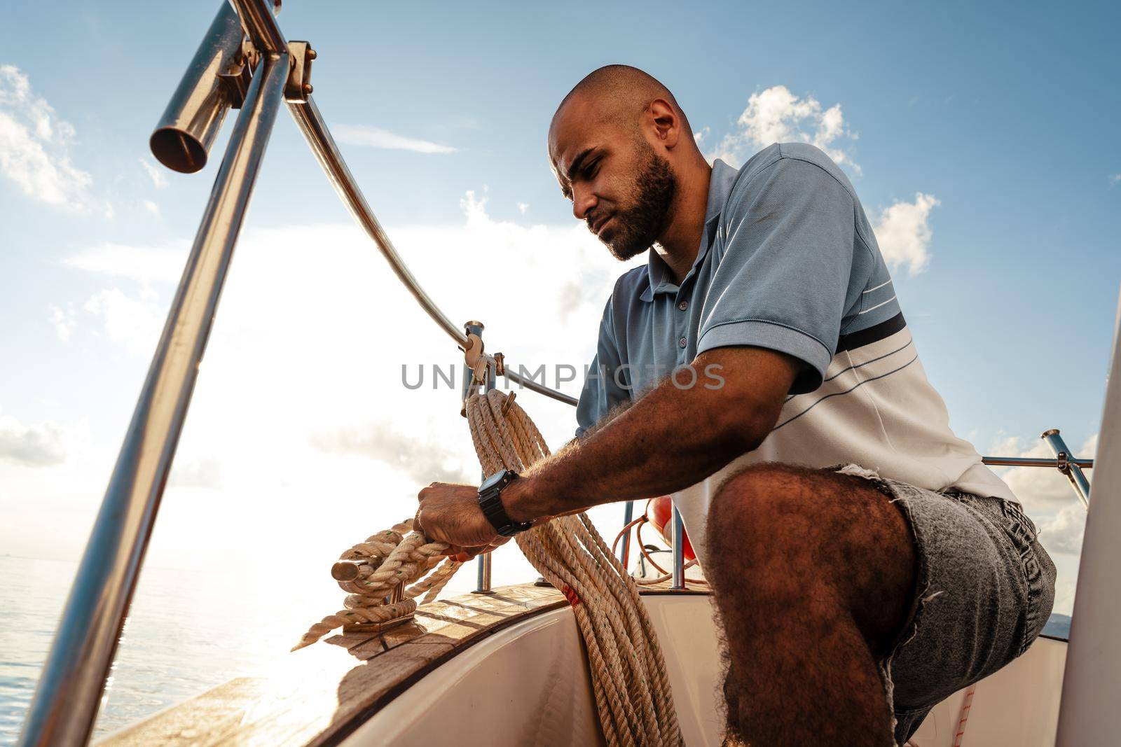 Young african american sailor tying ropes on sailboat in the sea on sunset, close up