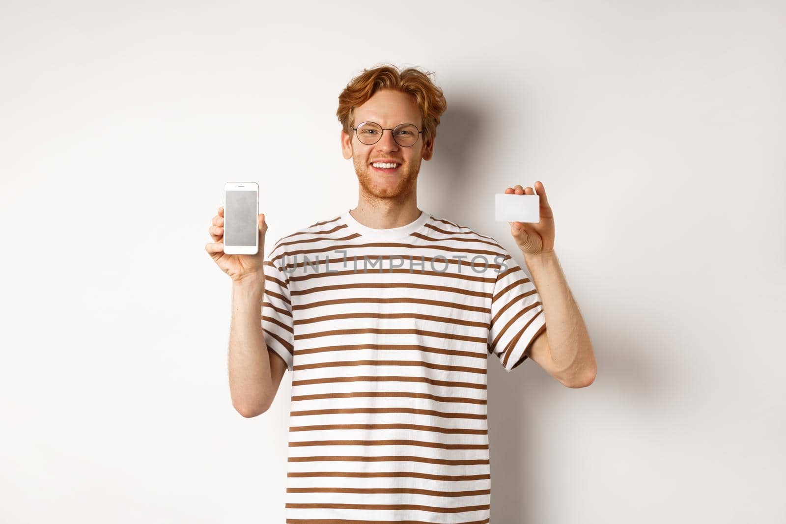 Shopping and finance concept. Young man showing blank mobile screen and plastic credit card, smiling at camera, white background by Benzoix