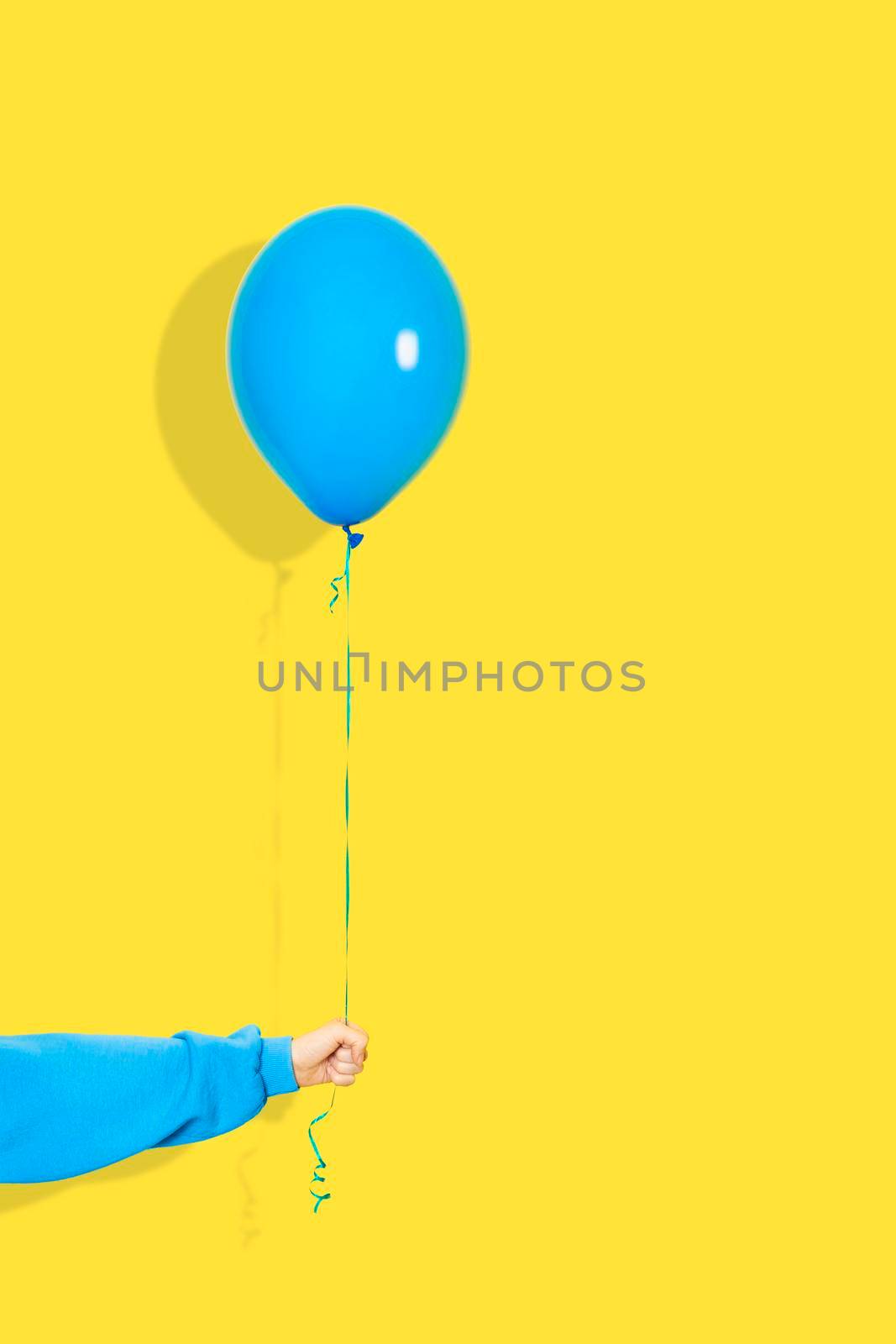 Female hand hold blue balloon. Party or present concept. Blue Balloon and hand in blue hoodie isolated on yellow backgroun by esvetleishaya