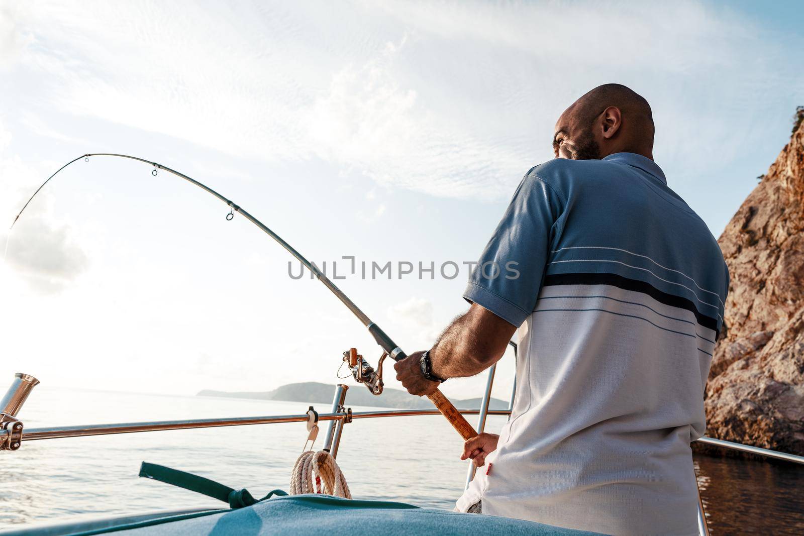 Young african american man standing with fishing rod on a sailboat fishing in open sea on sunset, close up