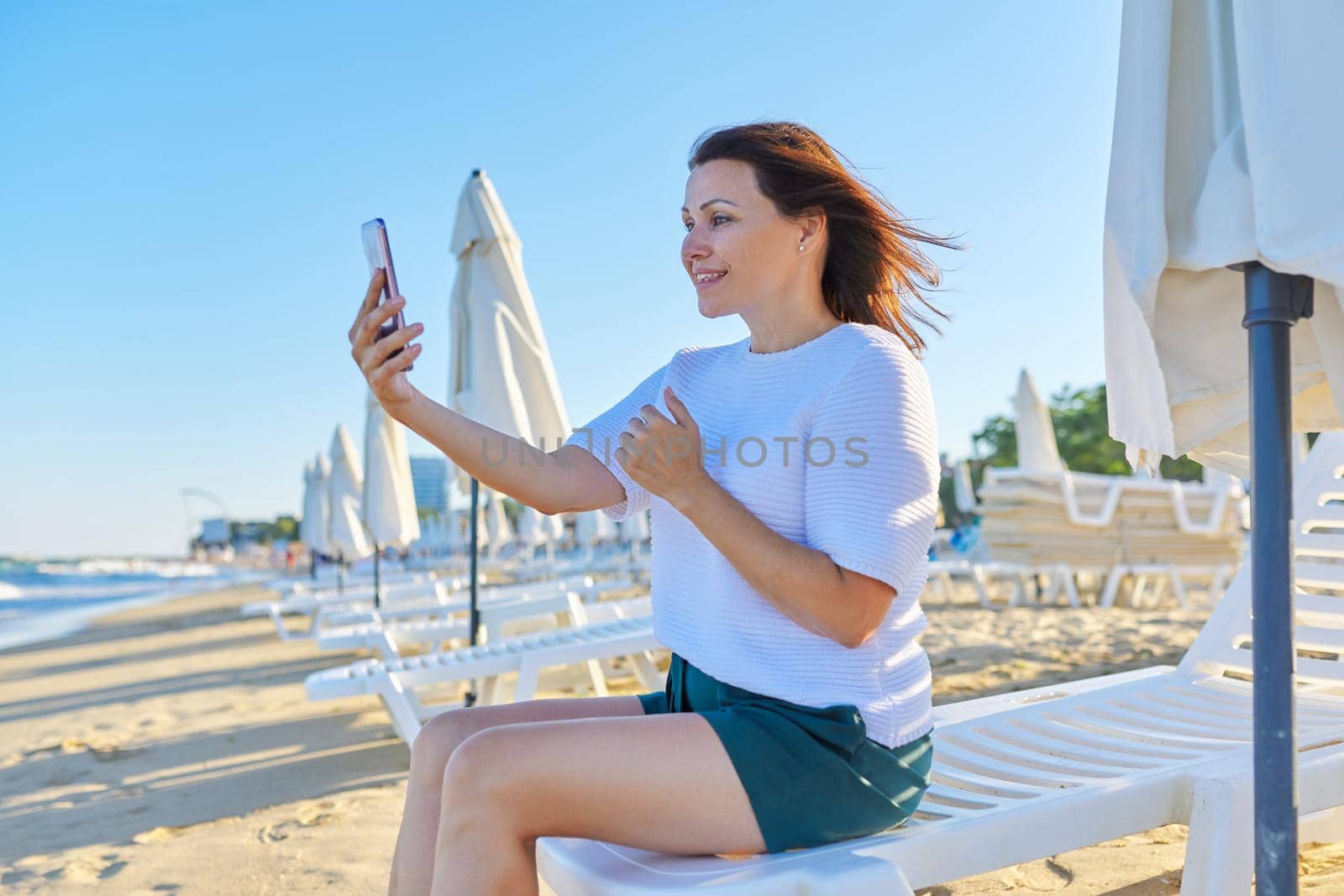 Happy middle-aged woman talking on smartphone using video call, female sitting in deck chair on sandy sea beach. Summer, vacation, weekend in nature, people of mature age