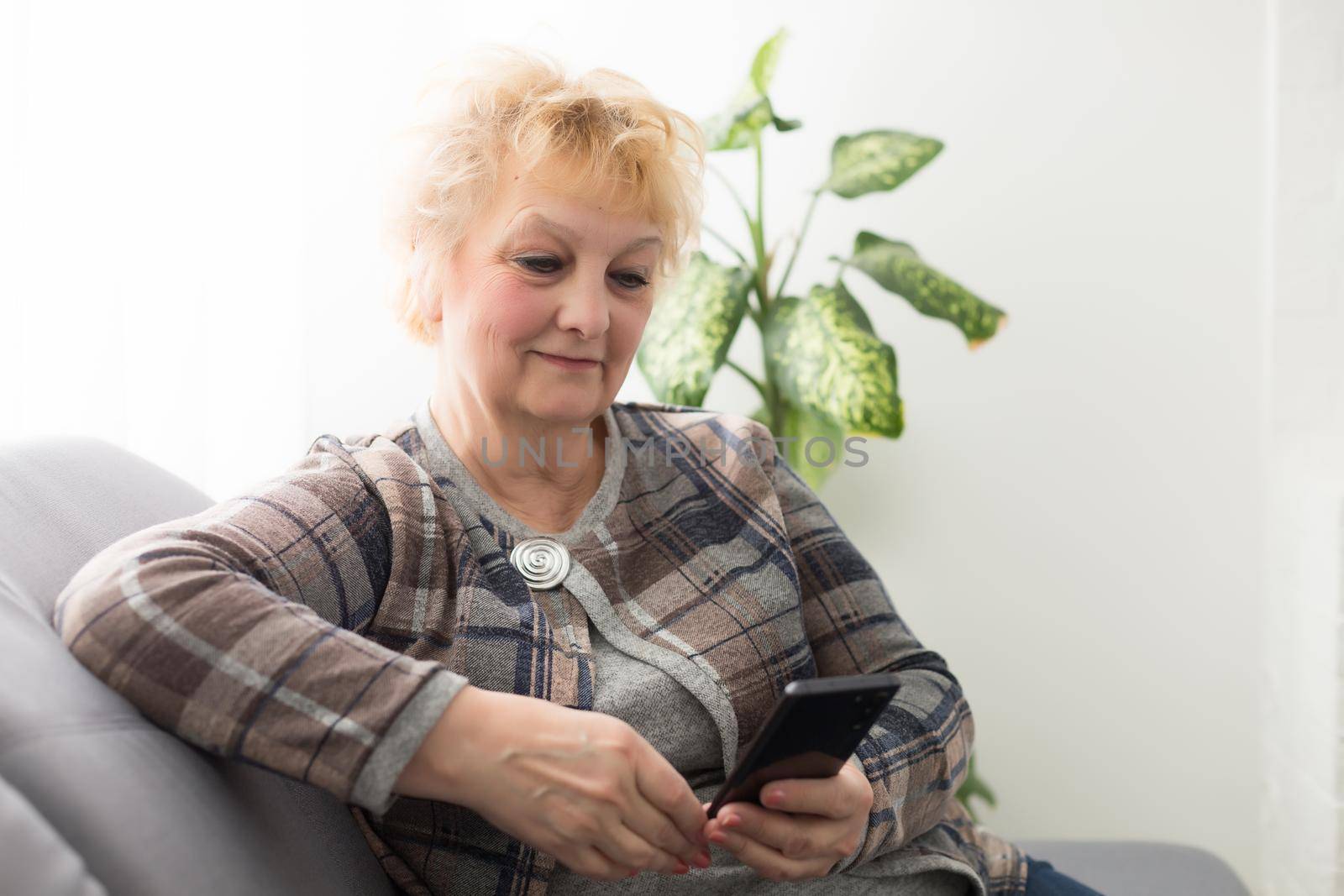 Senior woman with smartphone at home standing at the window