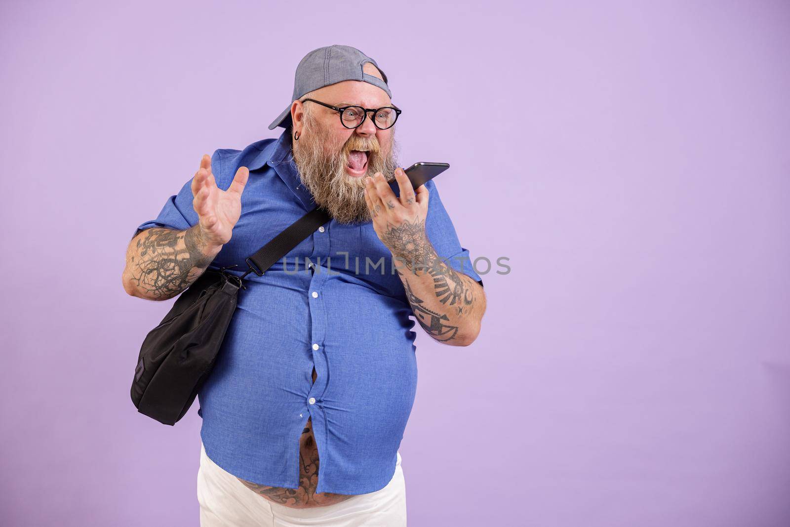 Angry emotional man with overweight yells by mobile phone using loudspeaker mode posing on purple background in studio