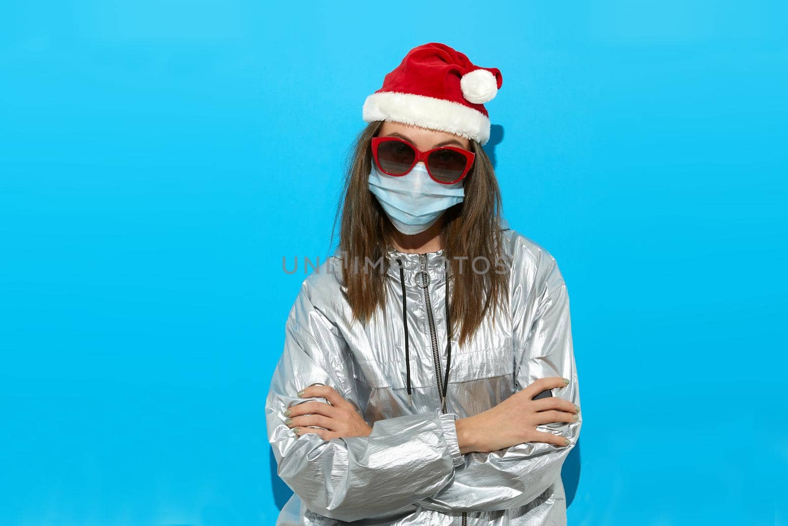 Serious female wearing Santa hat and protective mask and red sunglasses standing on blue background in studio and looking at camera Arms crossed