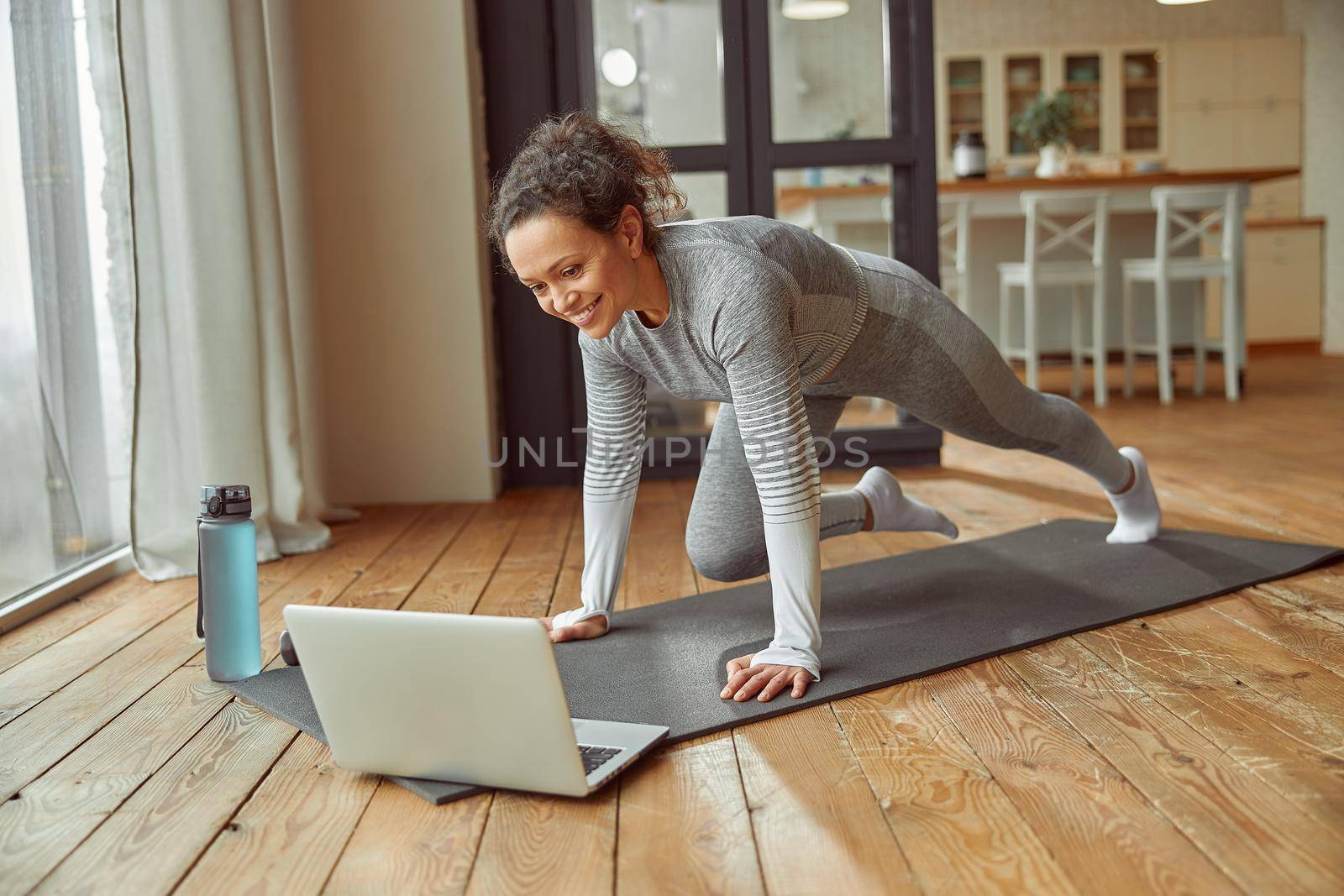 Cheerful slim woman is doing plank on mat while watching video on notebook at home