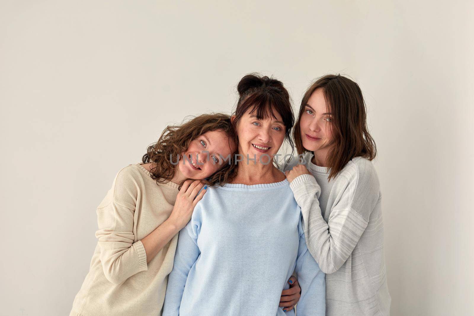 Cheerful adult mother embracing charming young daughters while standing on white background in studio and looking at camera