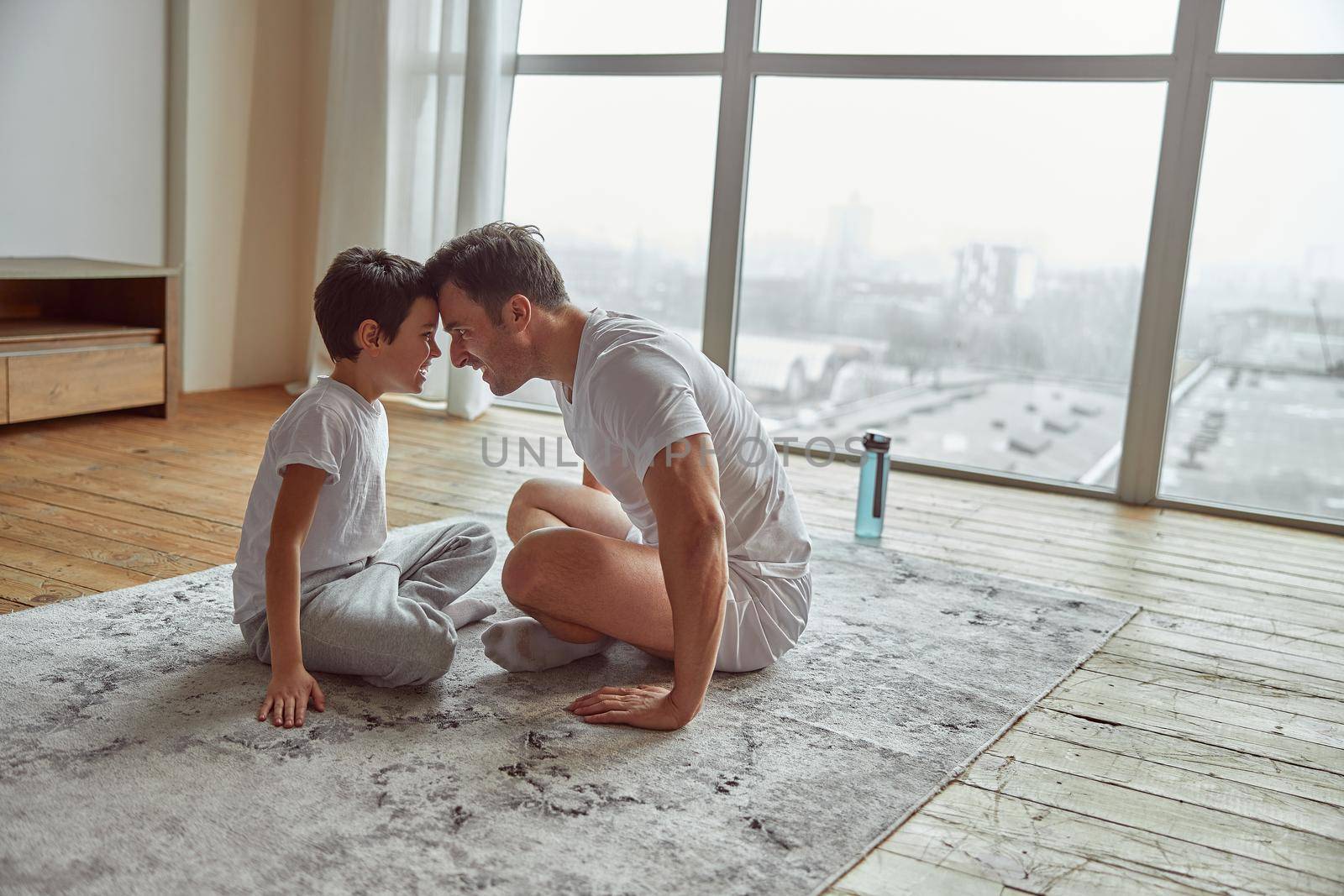 Cheerful father is sitting forehead to forehead to little boy while encouraging themselves for training at home