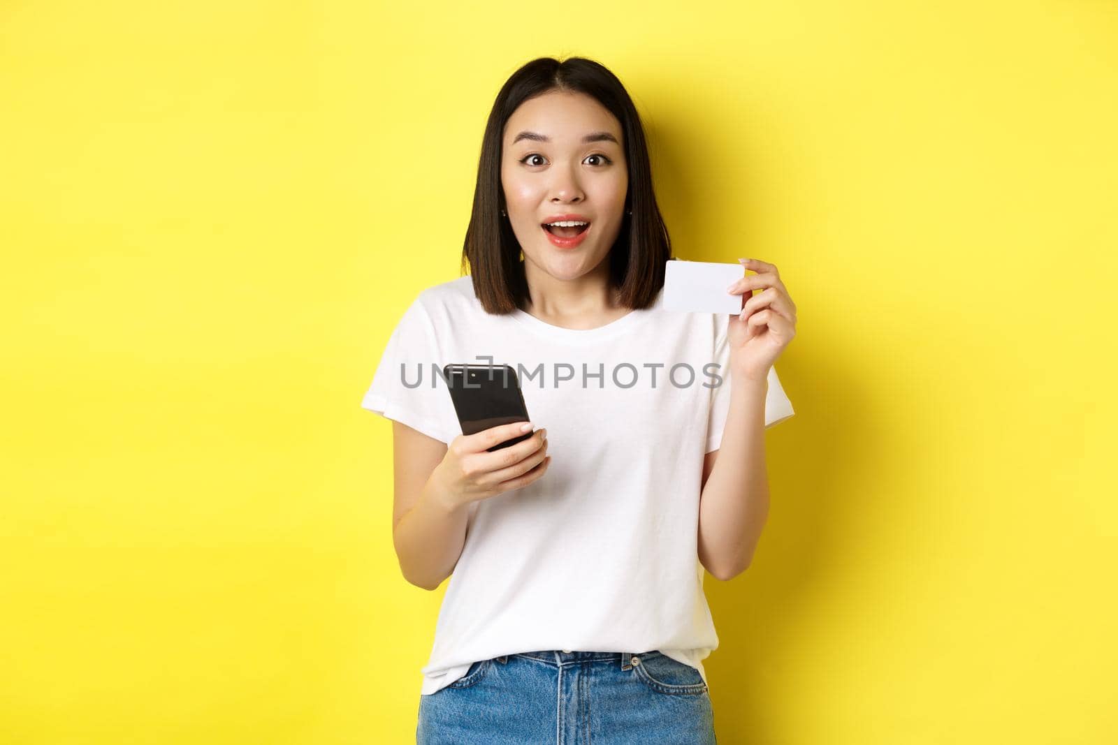 E-commerce and online shopping concept. Excited asian woman order in internet, holding smartphone and plastic credit card, standing over yellow background by Benzoix