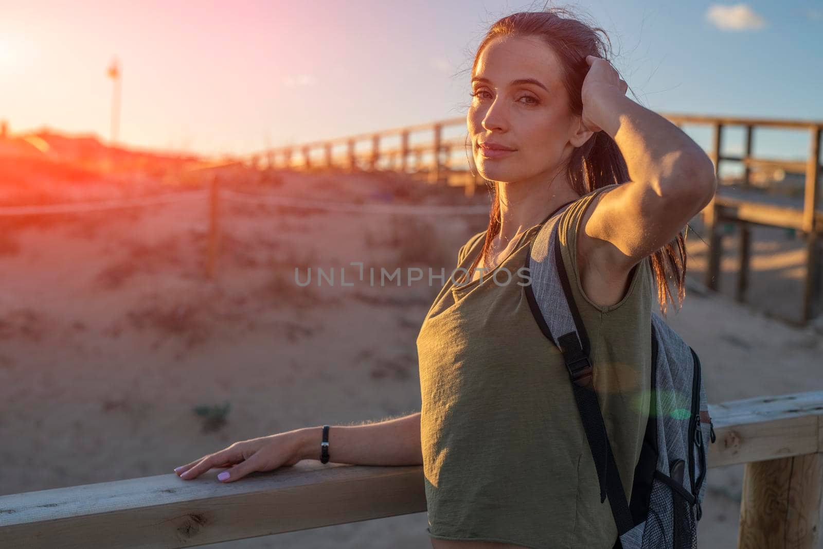 Fashionable slender young brunette girl with backpack posing on a background wooden walkways and sand beach at sunset.