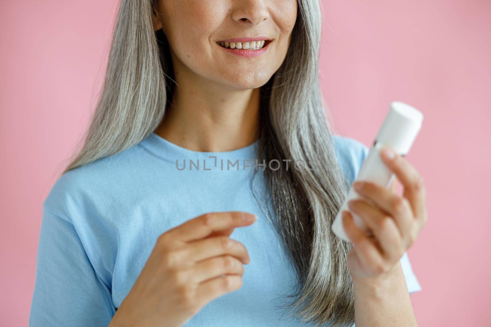 Smiling grey haired Asian lady holds bottle of cosmetic product on pink background in studio closeup. Mature beauty lifestyle