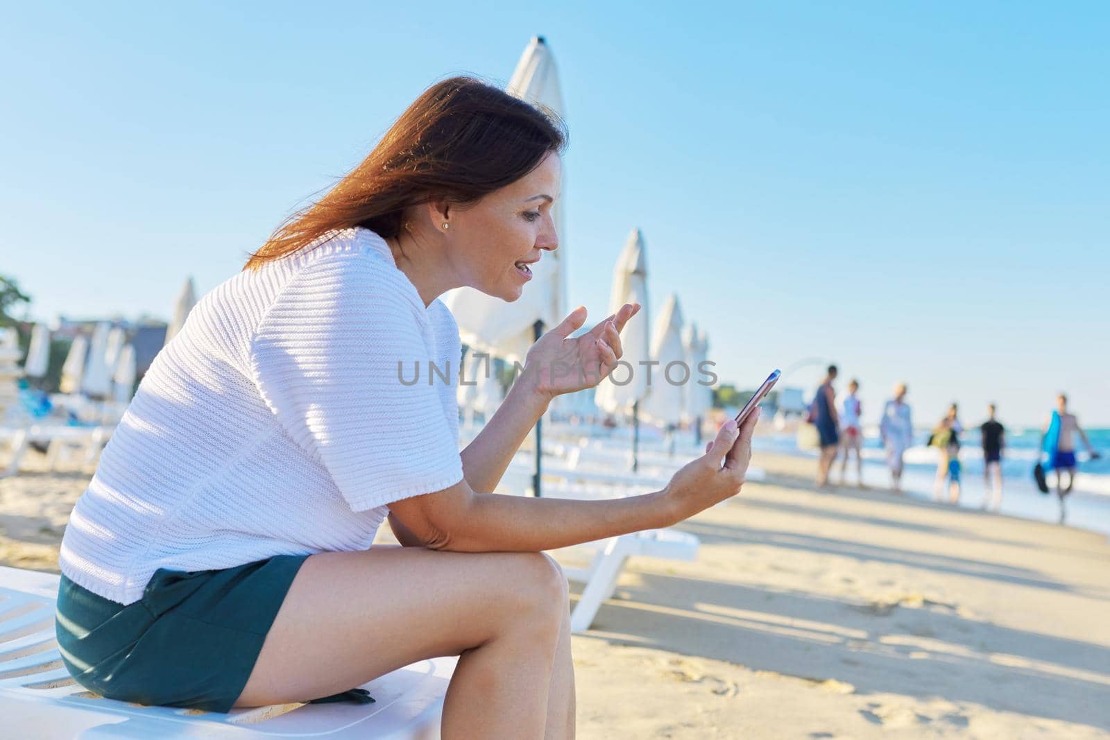 Happy middle-aged woman talking on smartphone using video call, on beach by VH-studio