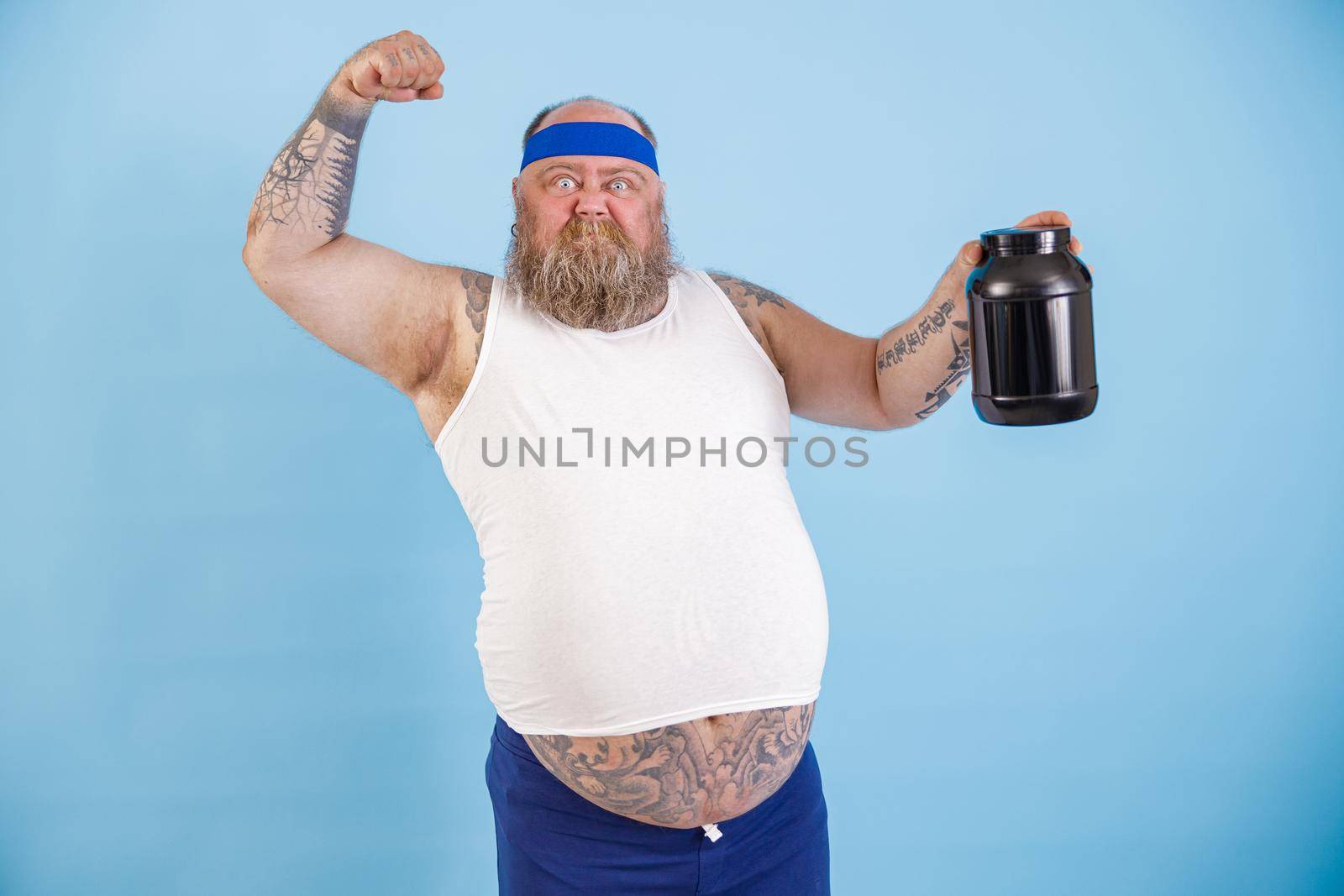 Strong bearded man with overweight shows muscles holding large bottle of sport nutrition standing on light blue background in studio