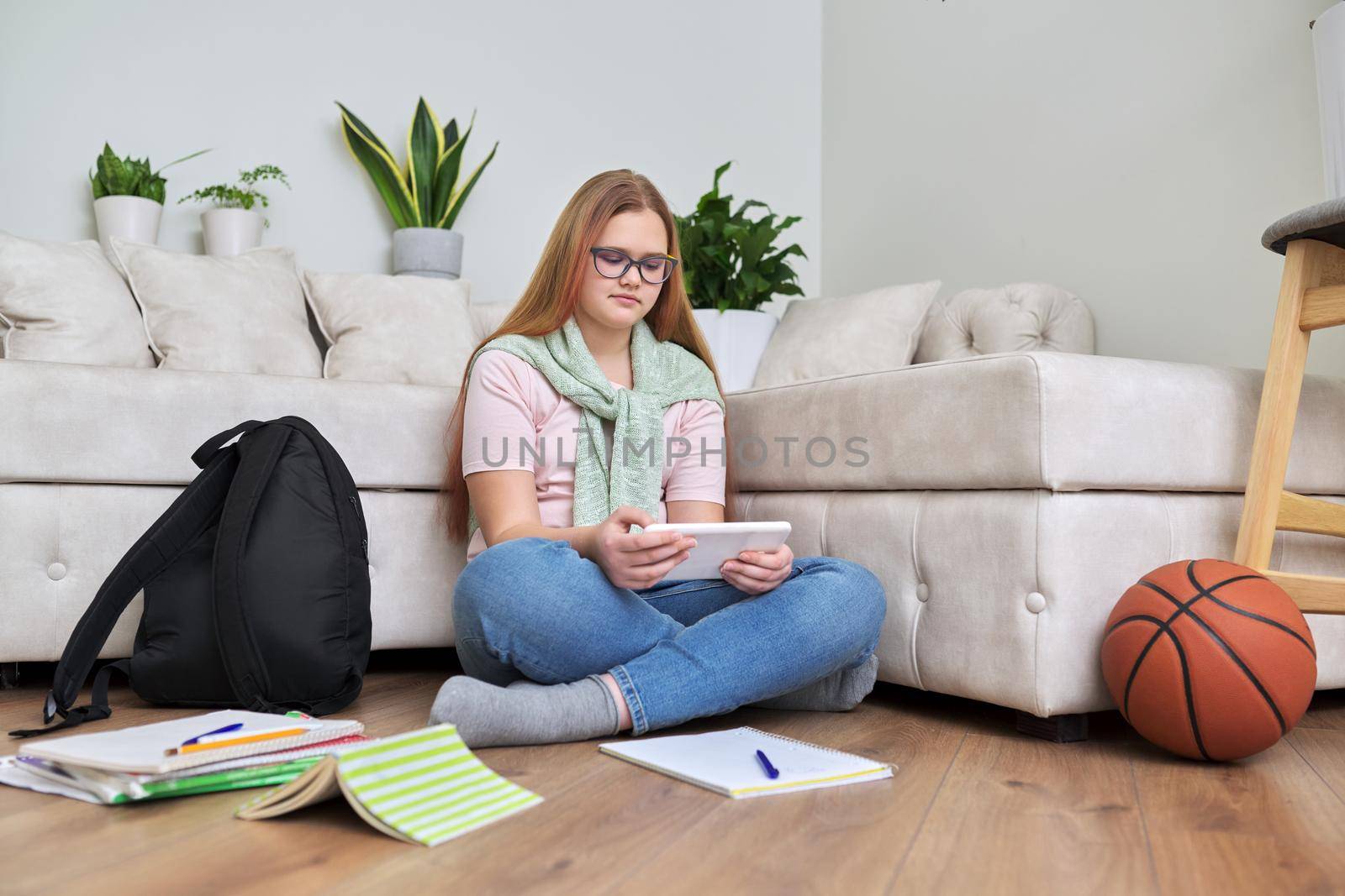 Portrait of teenage girl 12, 13 years old sitting at home on floor with school books, digital tablet. Online studies, e-education, resting schoolgirl using Internet modern digital technologies gadgets