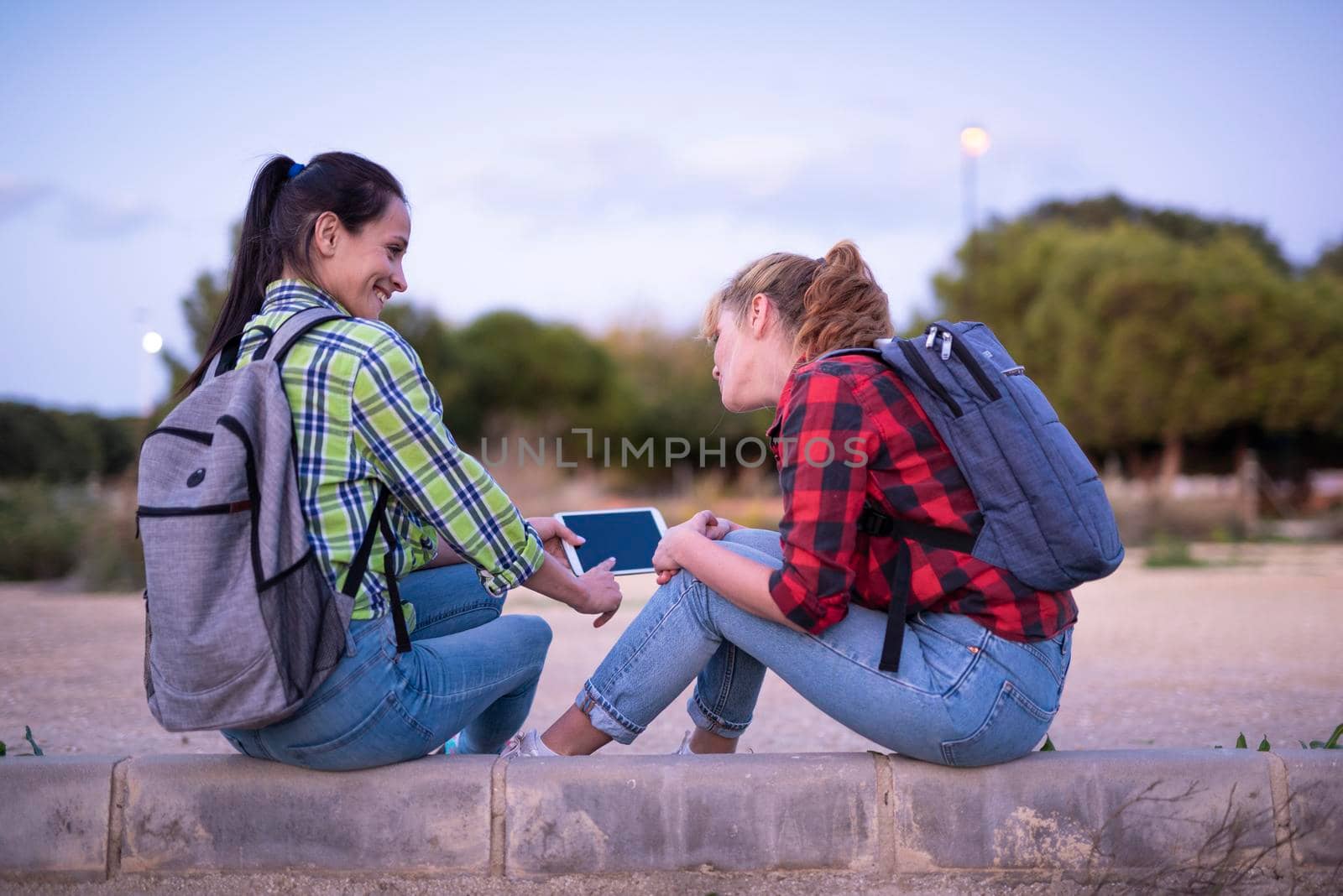 two young women students with backpack holding and looking at the tablet and smiling. Girl shows something on the screen. by Alexxoma