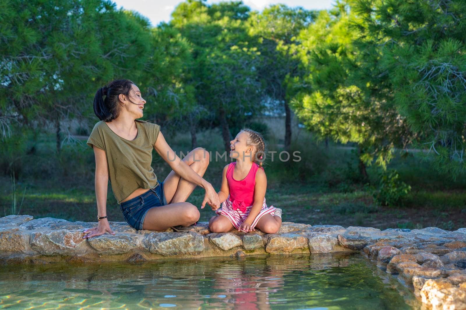 Beautiful mother and her little daughter outdoors. Beauty Mum and her Child playing with water in Park together at sunset. Outdoor Portrait of happy family. Mother's Day.