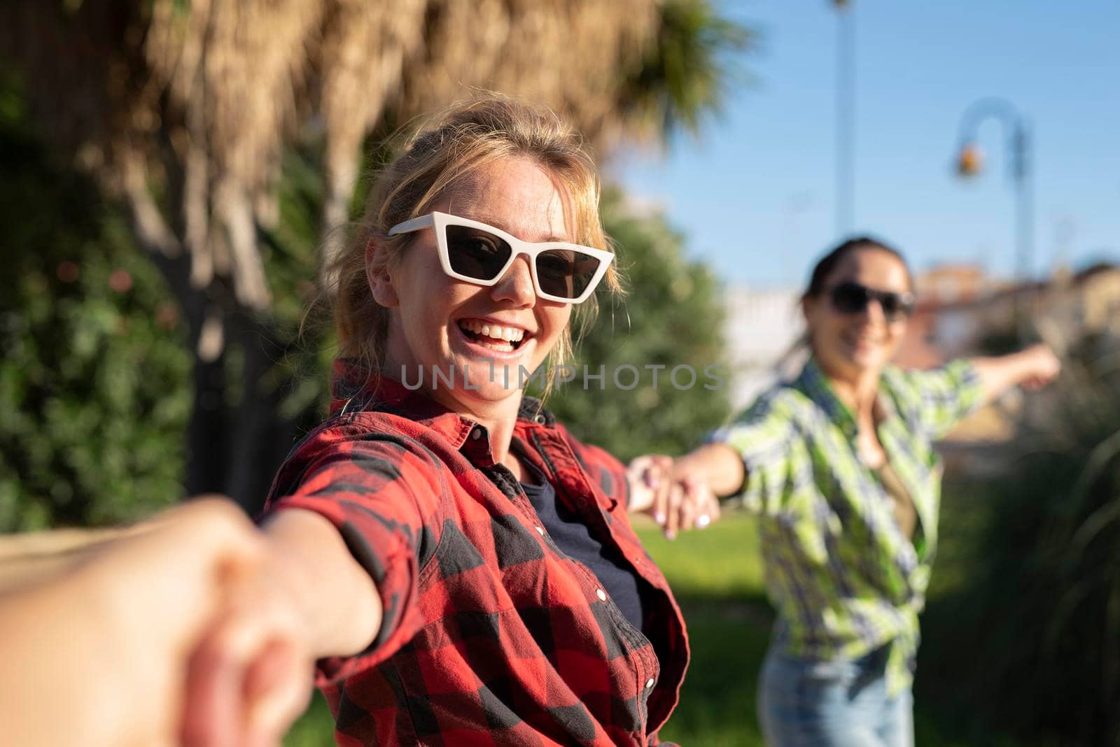 Pretty young caucasian woman in sunglasses pulling along by the hand invitingly with a happy smile as her friend on the background. Sunny summer day by Alexxoma