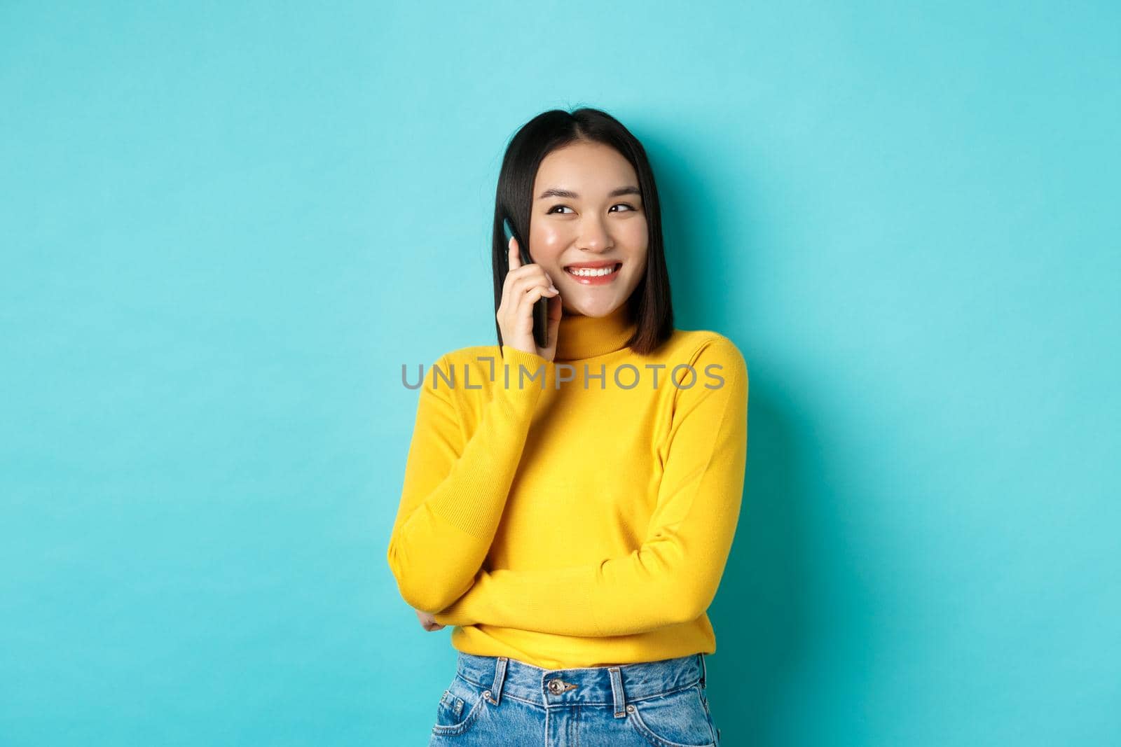 Attractive korean woman in yellow sweater, having conversation and smiling, talking on mobile phone, looking left at copy space, standing over blue background by Benzoix