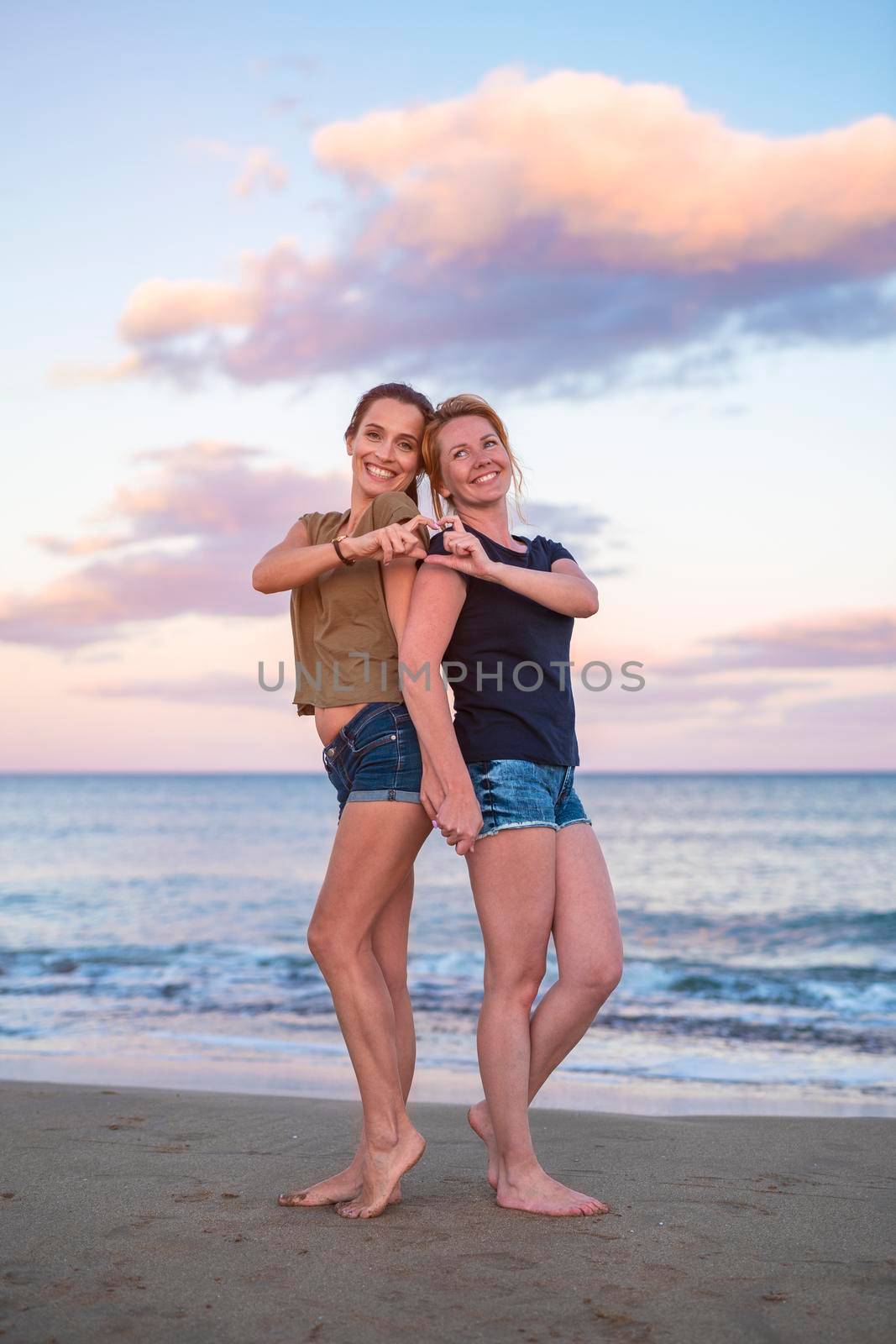 leisure and friendship concept - two happy smiling teenage girls or best friends at seaside making hand heart gesture at sunset.